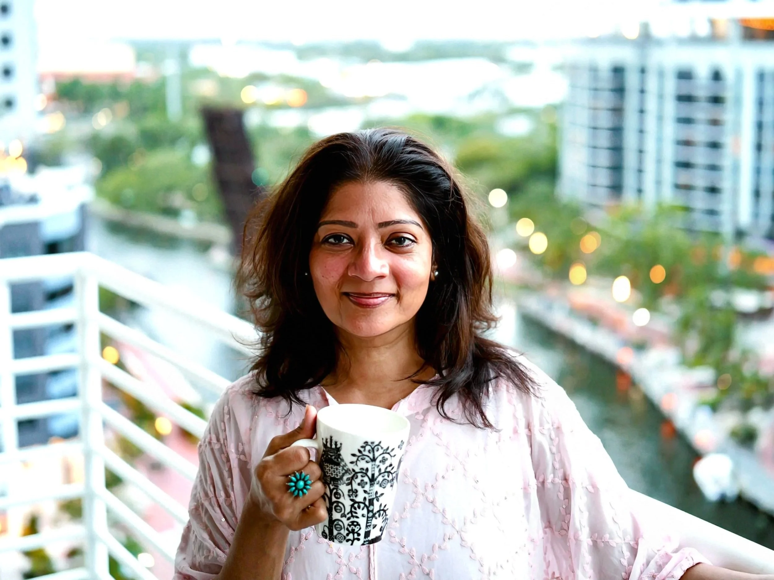 A woman with dark brown hair and earrings smiling while holding a white mug with black designs on a balcony during the evening. She is wearing a light pink top with textured details, and the background features a cityscape with high-rise buildings, lush greenery, and blurred lights.
