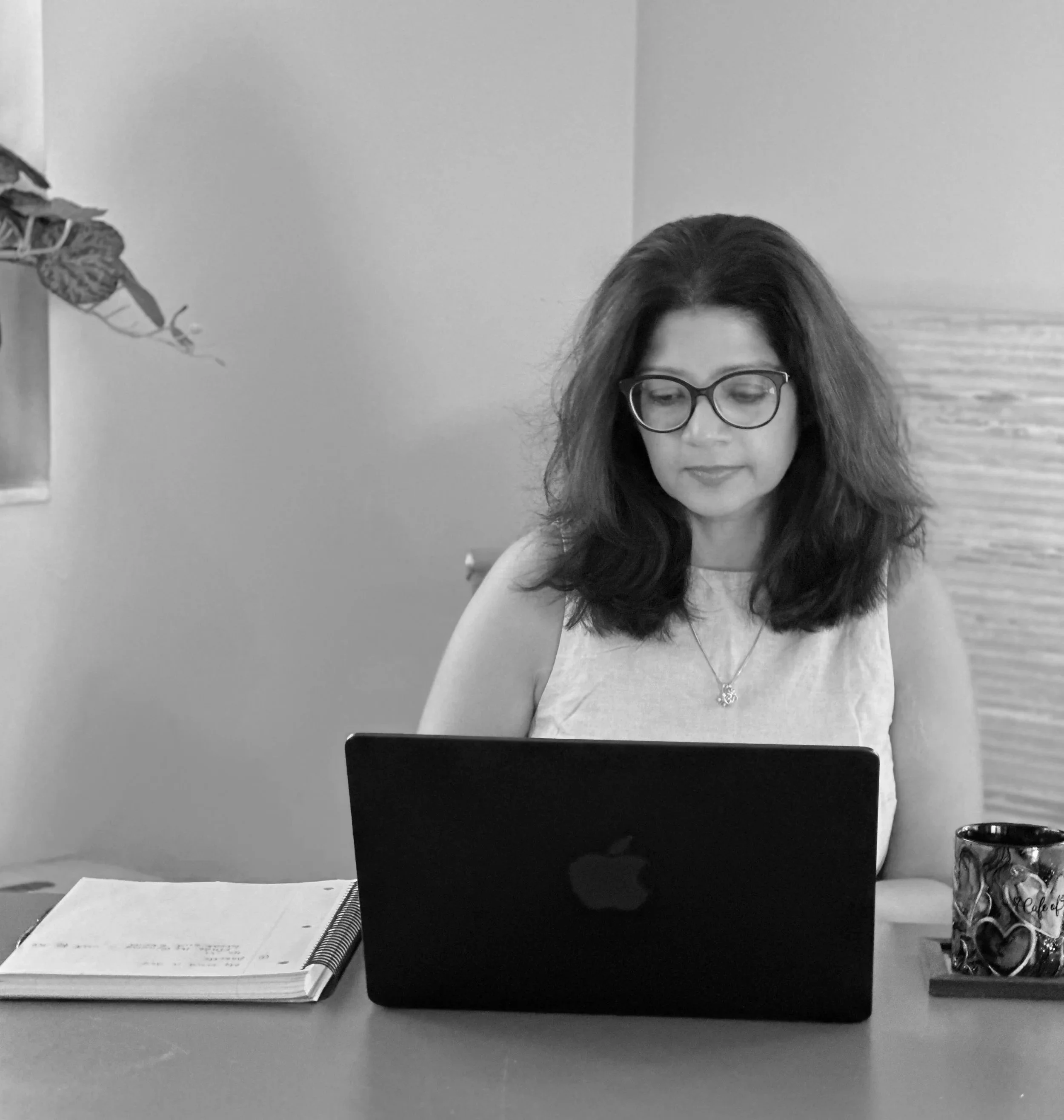 A woman with dark hair, glasses, wearing earrings and a necklace, sits at a desk with a laptop, an open notebook, and a coffee mug placed on the desk.