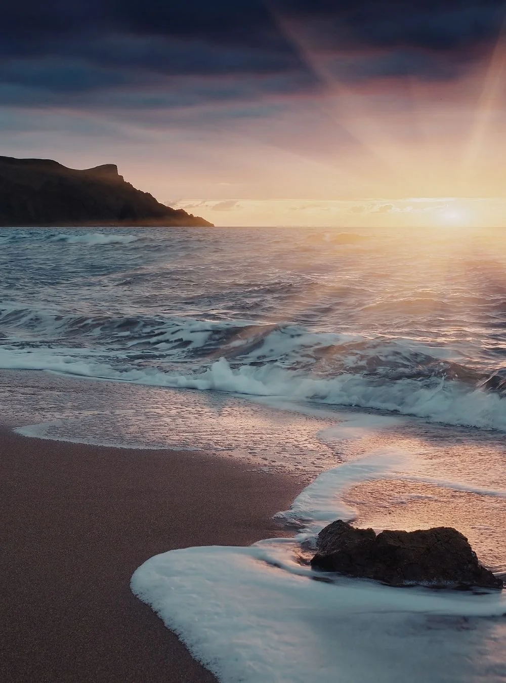 Sunset over ocean with waves crashing on the sandy beach and rocky shoreline, with cliffs in the distance under a partly cloudy sky.
