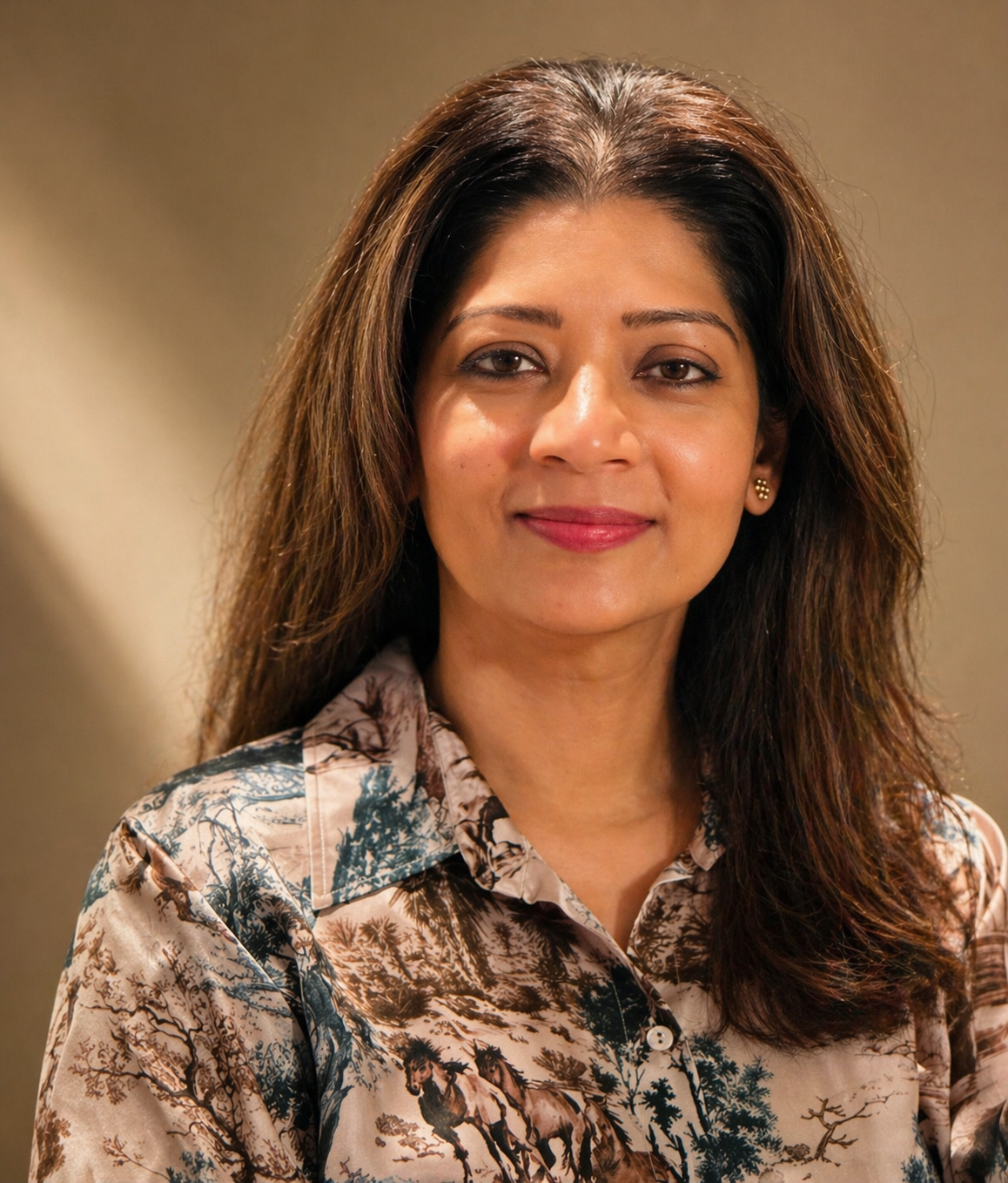 A woman with long dark hair and brown eyes, smiling, wearing a beige blouse with a nature scene print, standing in front of a neutral background.