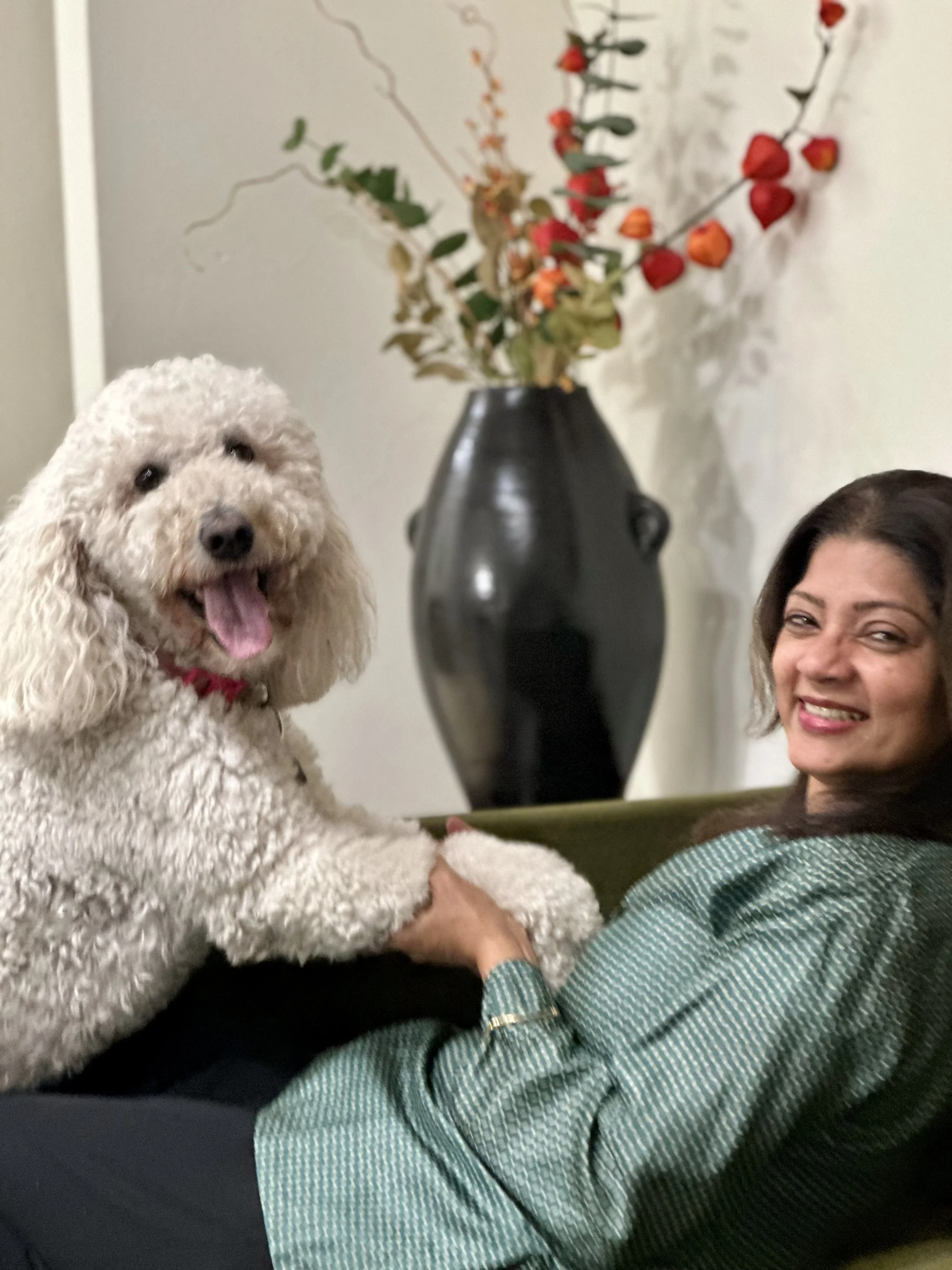 A woman holding a white curly-haired dog with a red collar, sitting on a couch. Behind them, a tall black vase with a dried floral arrangement featuring red and orange berries and green leaves. The woman is smiling and wearing a green and white patterned shirt.
