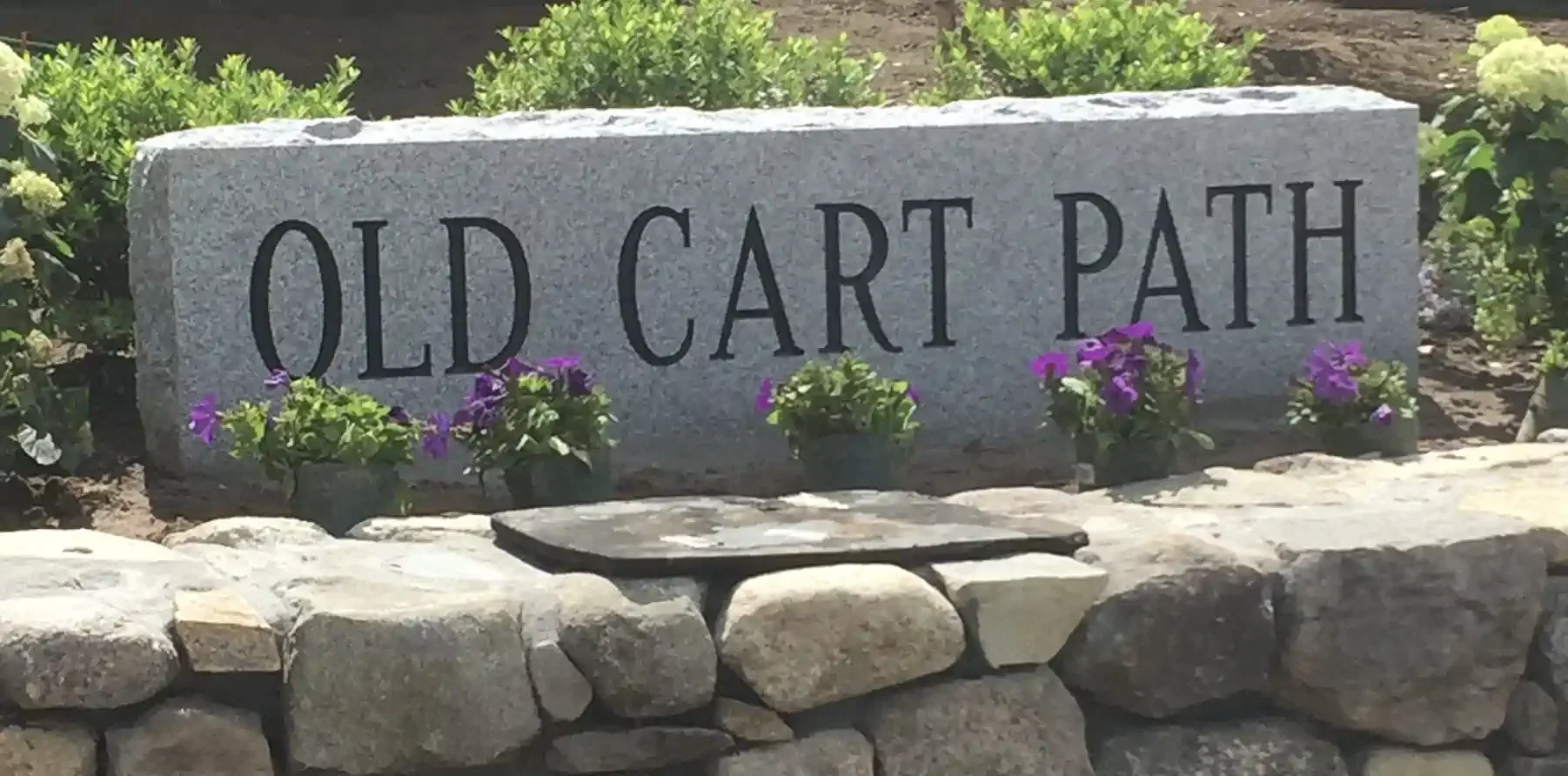 Stone sign reading 'Old Cart Path' surrounded by purple flowers and green bushes, with a stone border and a small table in front.