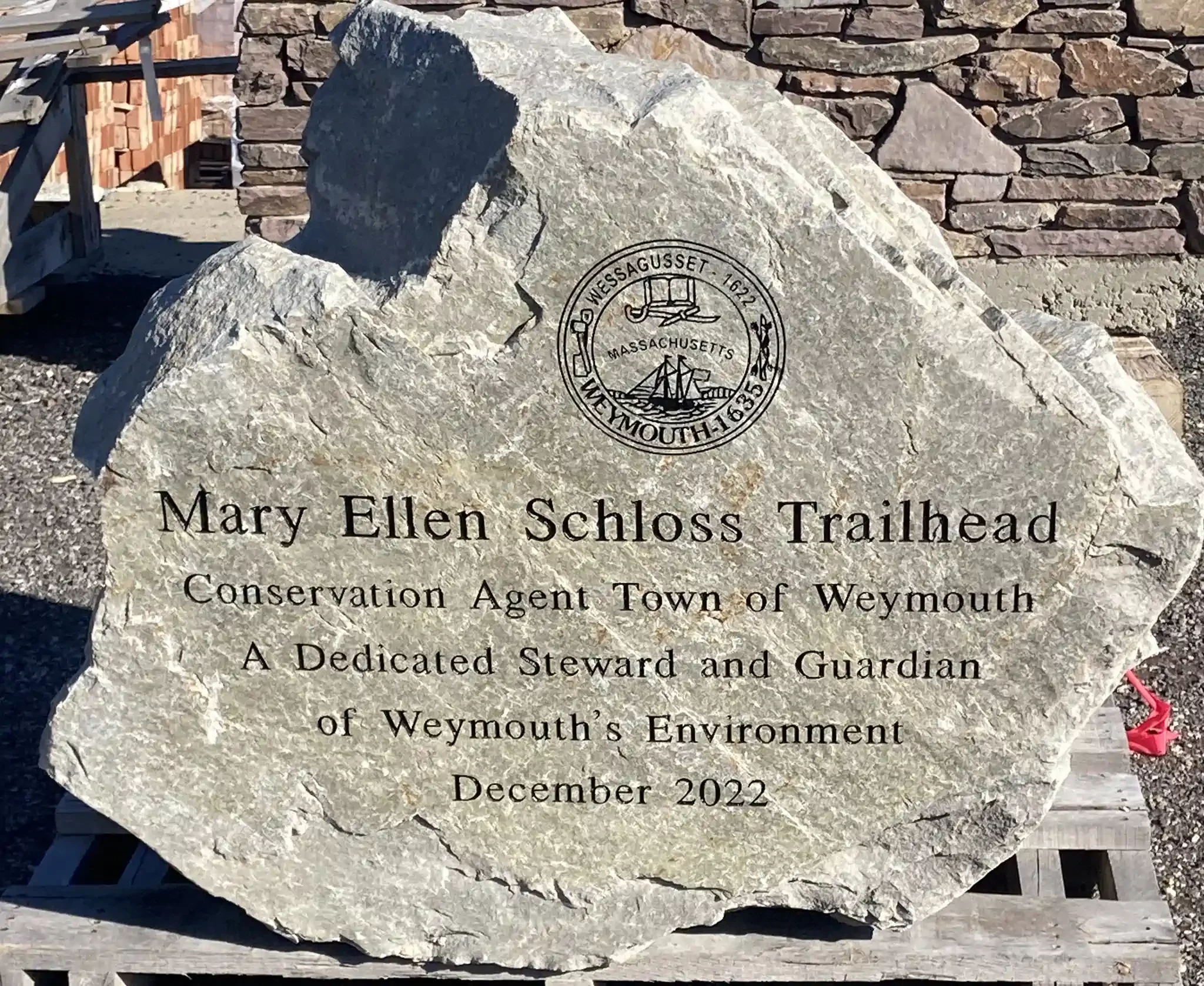 A granite memorial plaque with inscriptions and a circular emblem, placed outside against a stone wall.