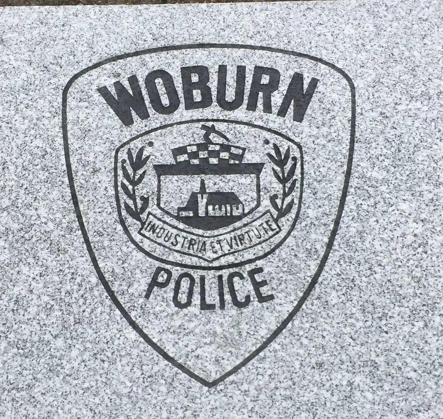 Black police badge with the words "Woburn Police" and a small illustration of a church surrounded by leaves and the phrase "Industria Tutte" on a speckled gray background.