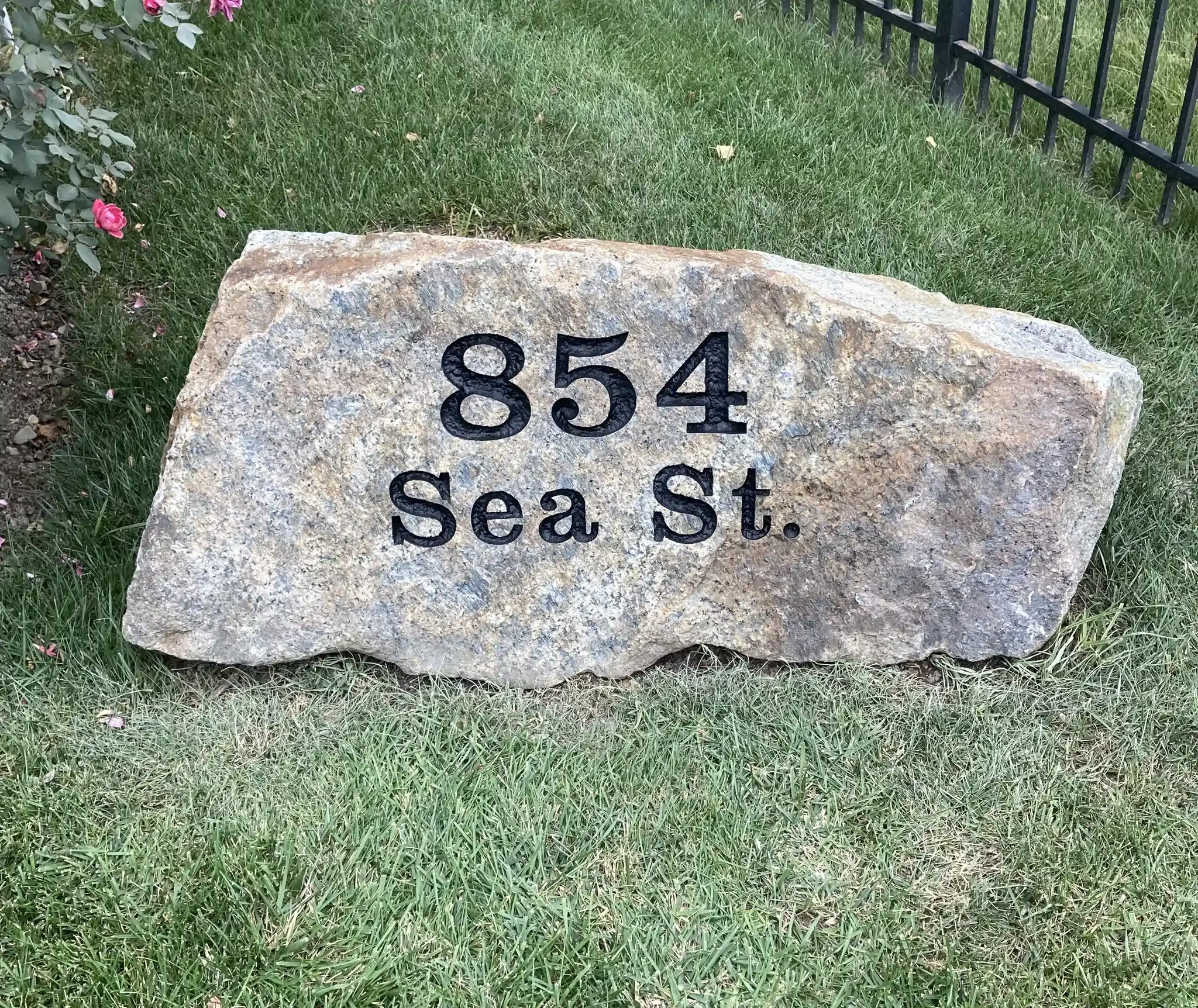 A large address stone with the number 854, situated in a grassy area with small pink flowers and a black metal fence nearby.