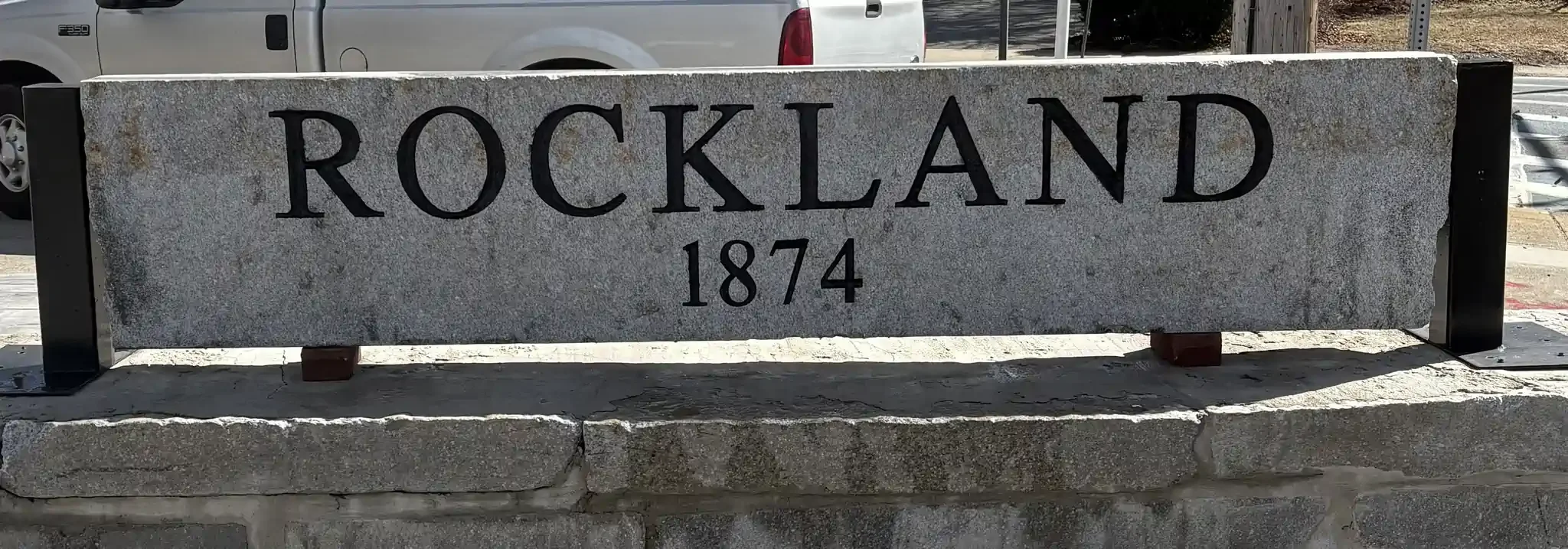 A gray stone sign with the word "Rockland" and the year "1874" engraved on it, set on a stone base in an outdoor setting.