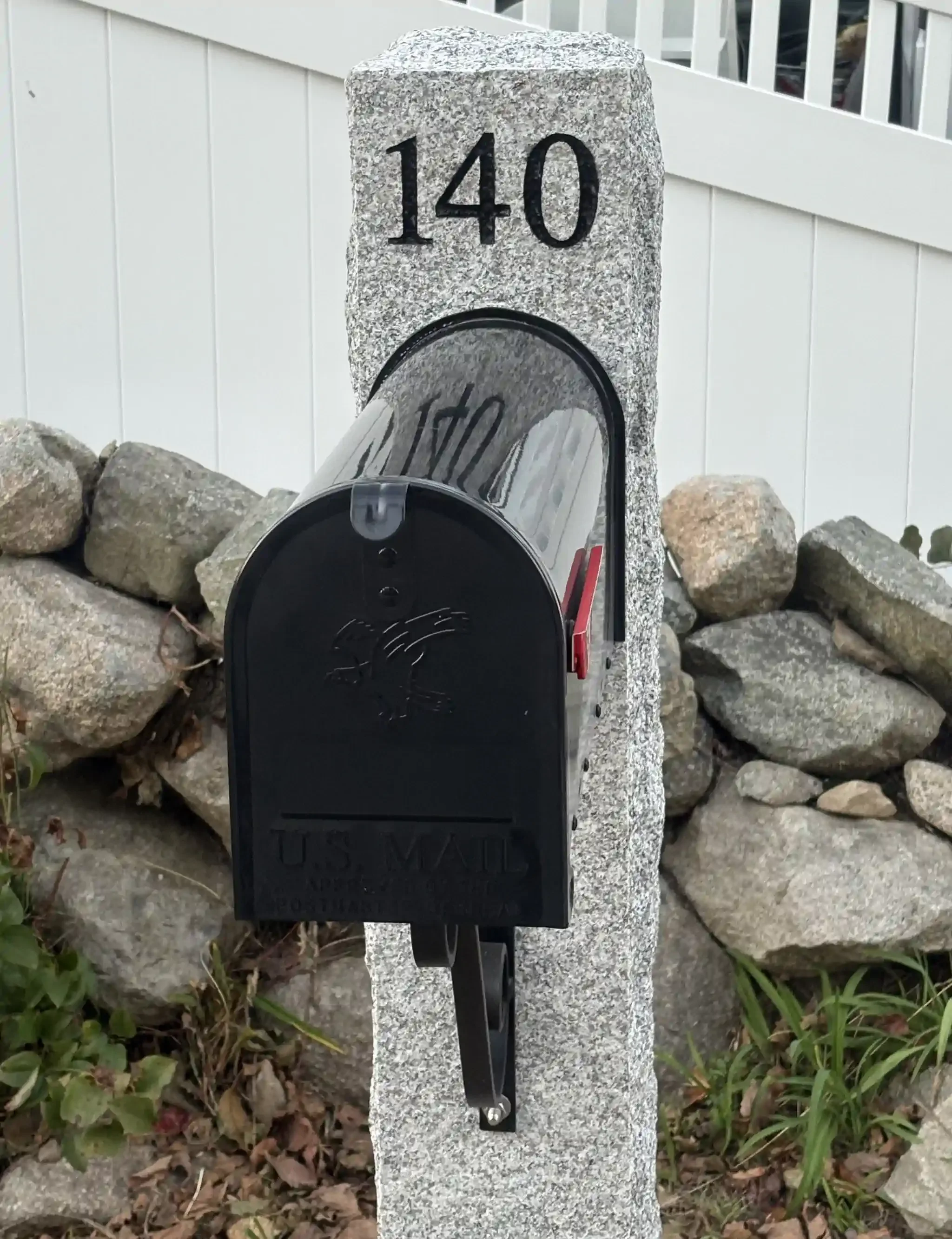 A black mailbox attached to a gray granite post with the number 140 engraved into it, surrounded by rocks and greenery.