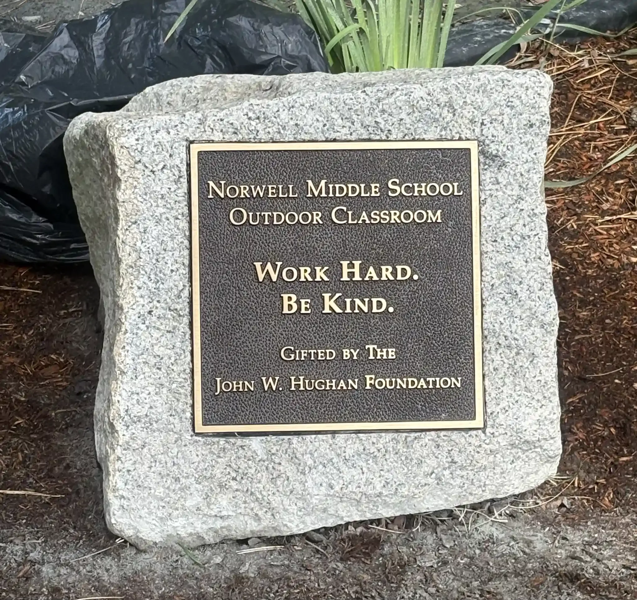 A plaque with the inscription 'Norwell Middle School Outdoor Classroom Work Hard Be Kind,' located outdoors amongst soil and plants.