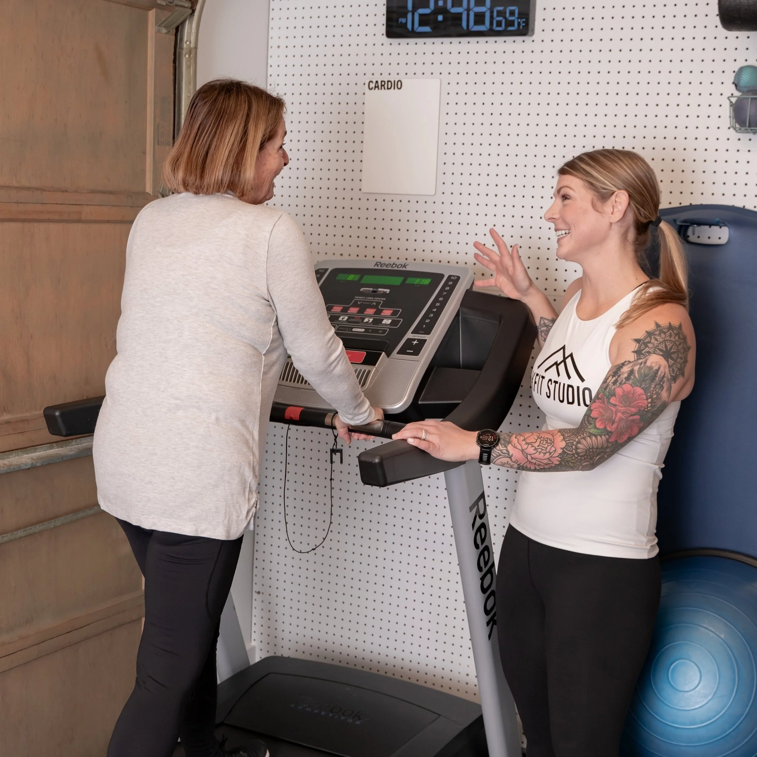 Two women talking near a treadmill in a gym, one using the treadmill and the other standing beside her.
