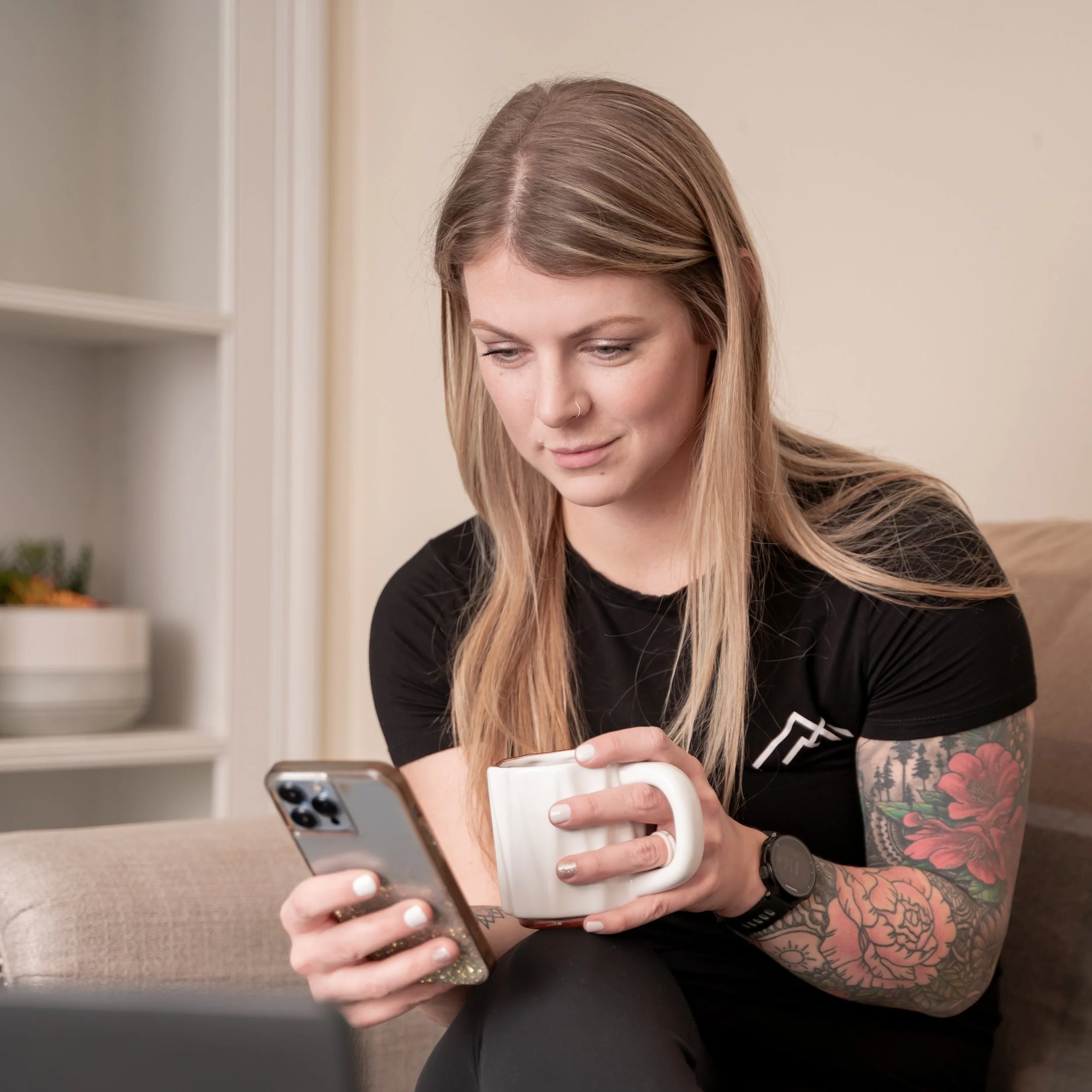 A woman with long blonde hair and tattoos on her right arm, sitting on a beige couch, looking at her phone and holding a white mug.
