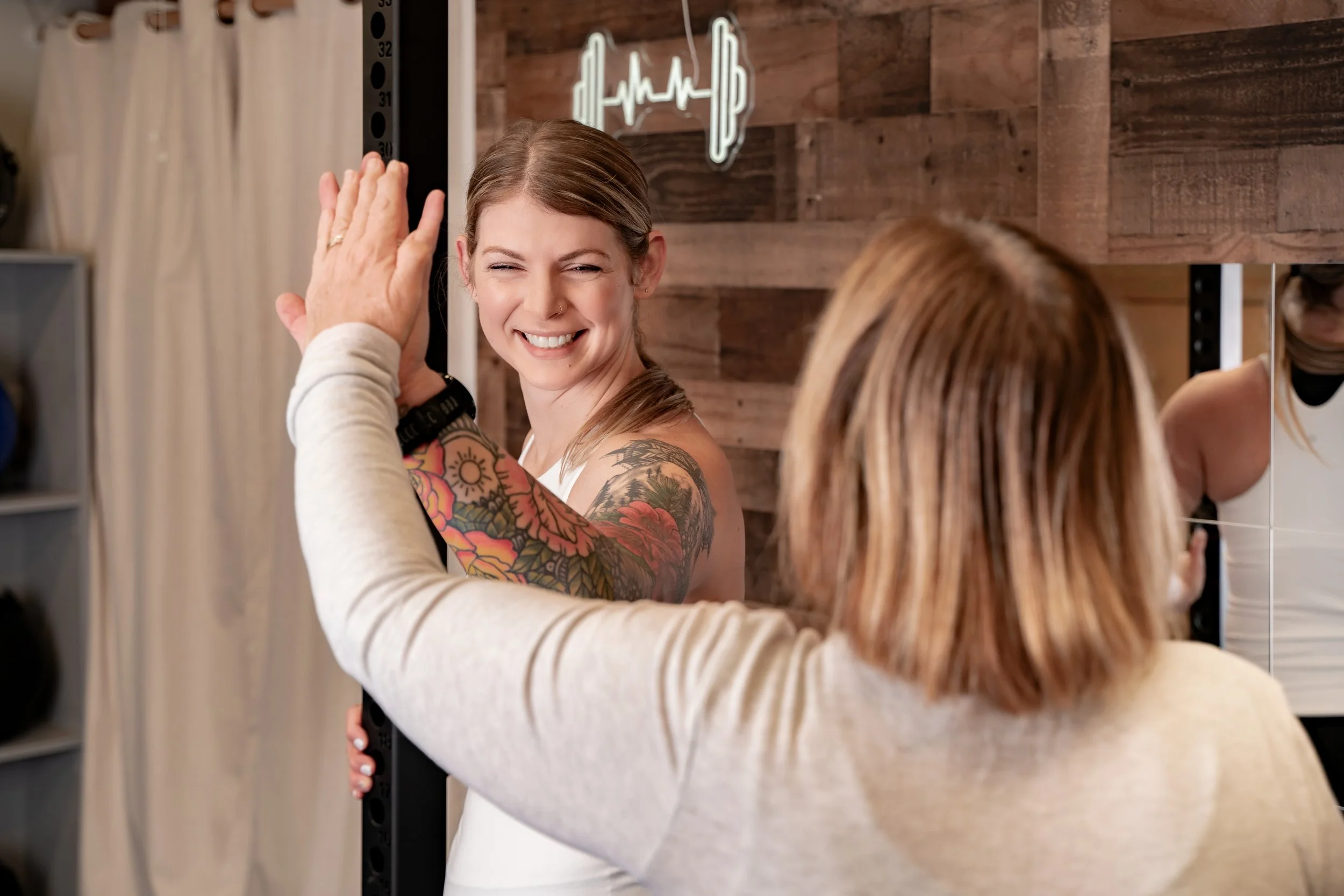 A woman with tattoos and a big smile gives a high five to another woman with shoulder-length red hair in a gym or fitness studio.