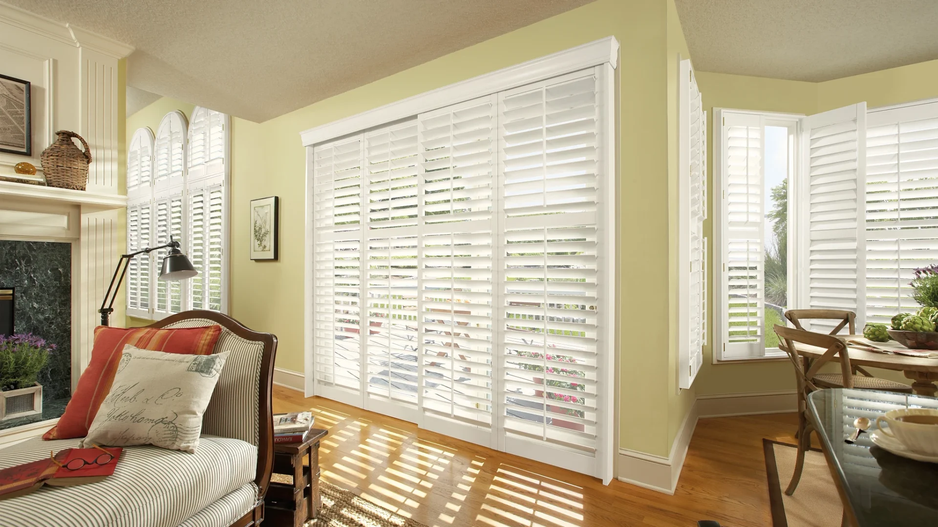 Living room with large white plantation shutters, a cushioned chair with striped fabric, a lamp, and a small table with books and glasses.