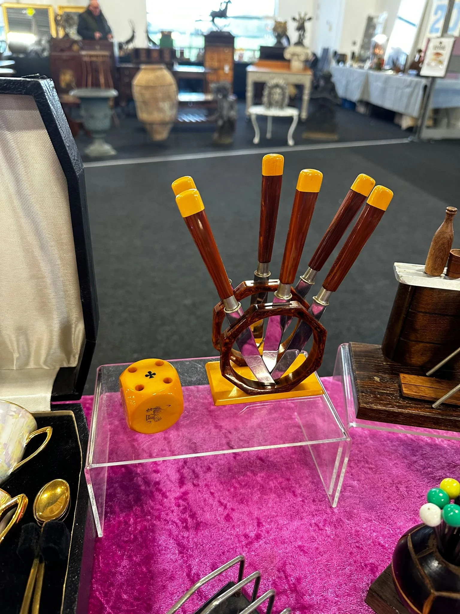Vintage Catalin Handled Knives with Dice Display
A display of vintage knives featuring brown Catalin handles with yellow Catalin tips, accompanied by a yellow Catalin die showing the number four. Photographed in an antique shop or auction setting, th