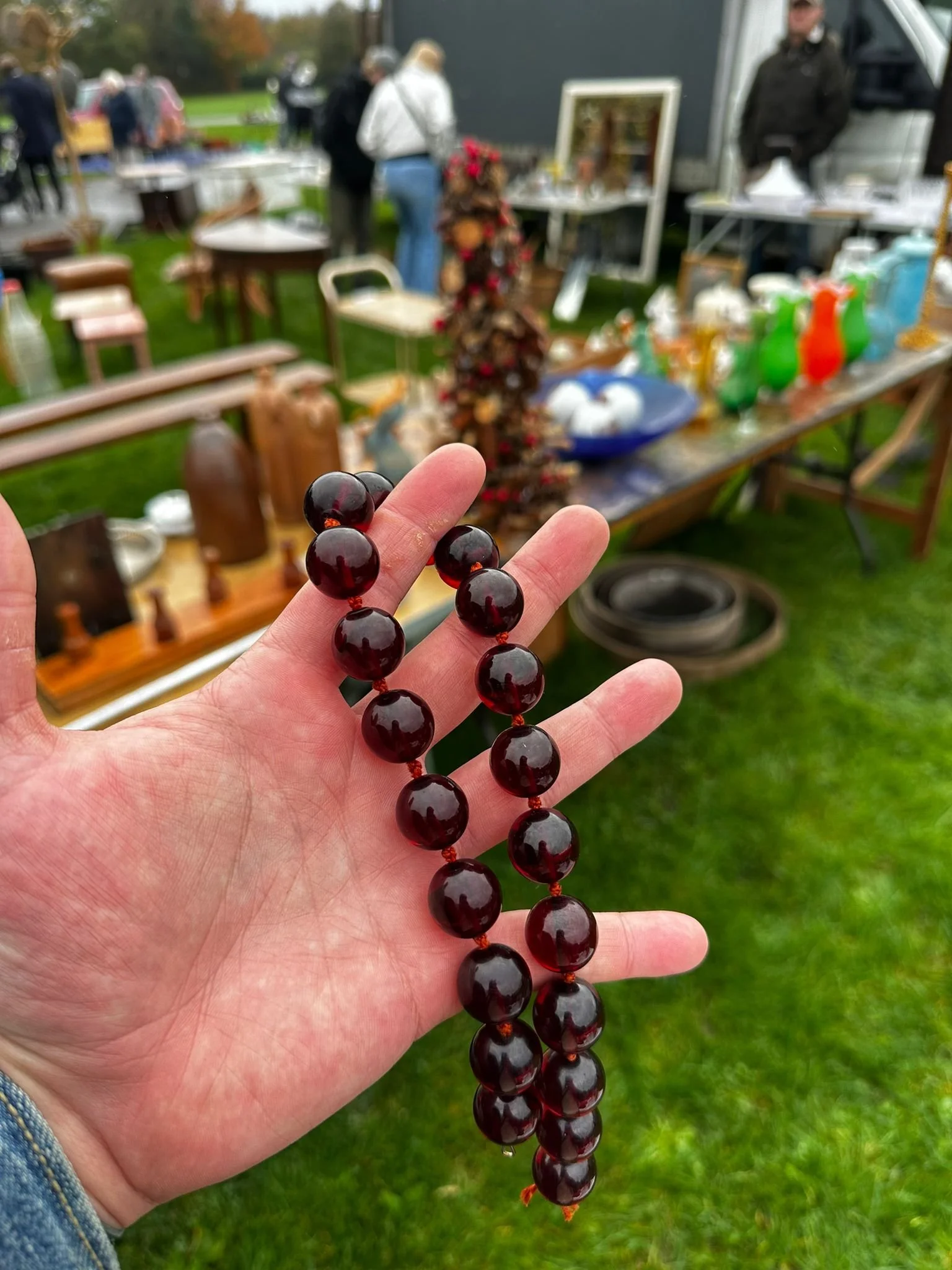 Vintage Cherry Amber Bead Necklace
A hand holding a string of dark red cherry amber beads, set against the backdrop of an outdoor flea market with tables of antiques and people browsing. The rich, deep tones of the beads capture the classic look and