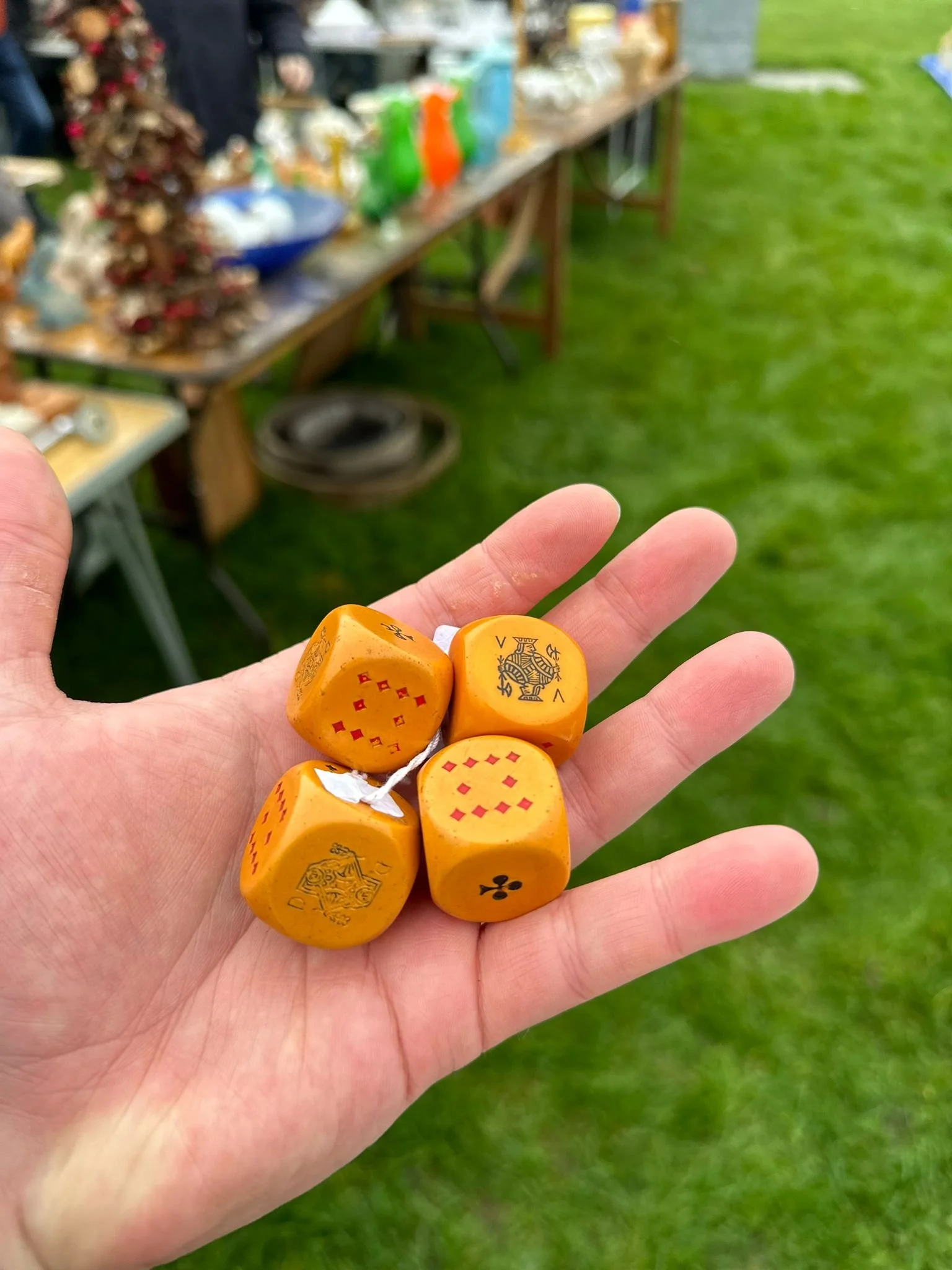 Vintage Catalin Bakelite Dice
A person’s hand holding four orange Catalin Bakelite dice, each decorated with red and black symbols including a club, a star, and checkered patterns. Photographed at an outdoor market with blurred tables of antiques in
