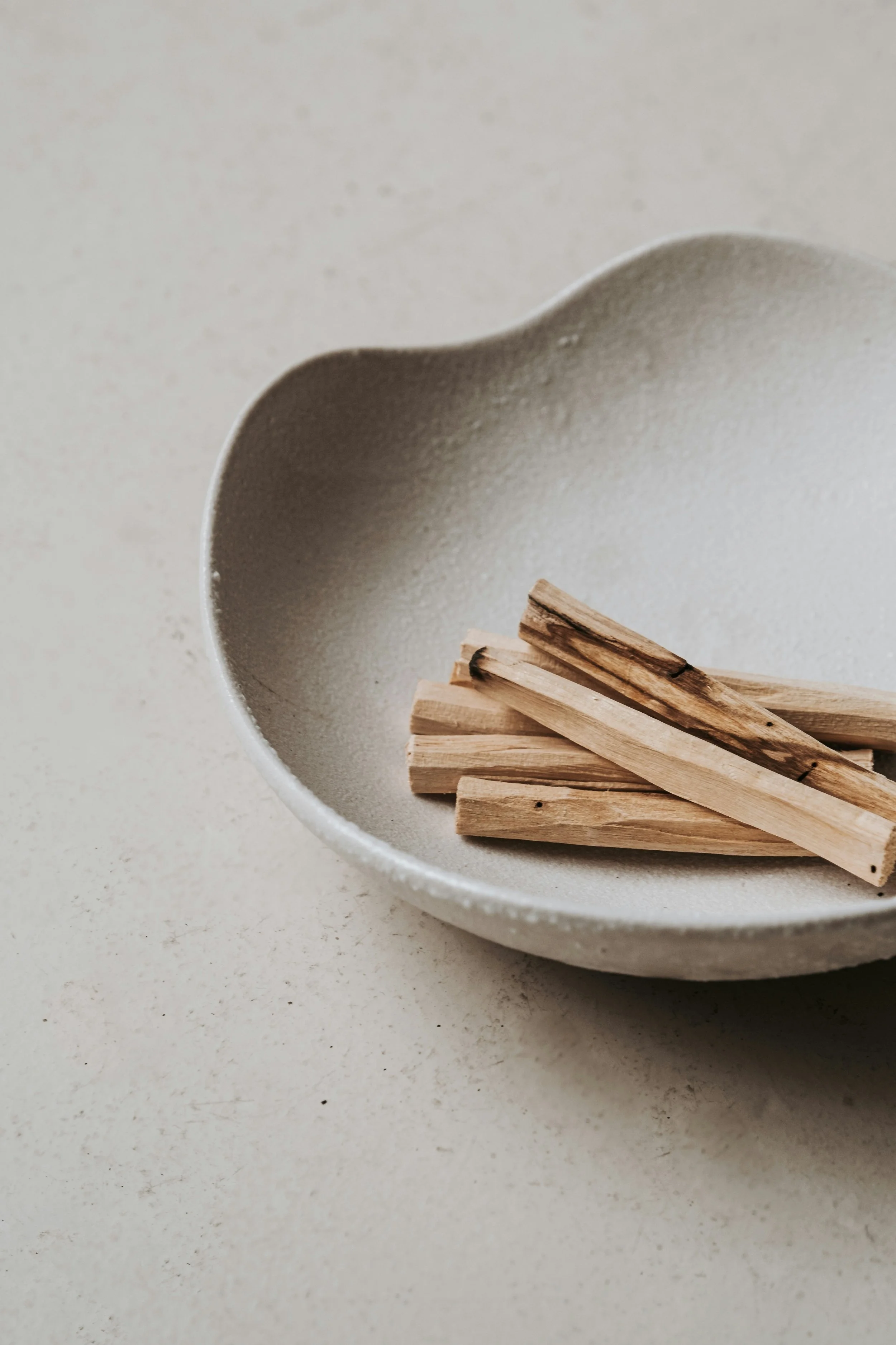 A close-up of a white, textured ceramic bowl with an irregular outer edge, containing several light-colored, charred and unburnt wooden sticks.