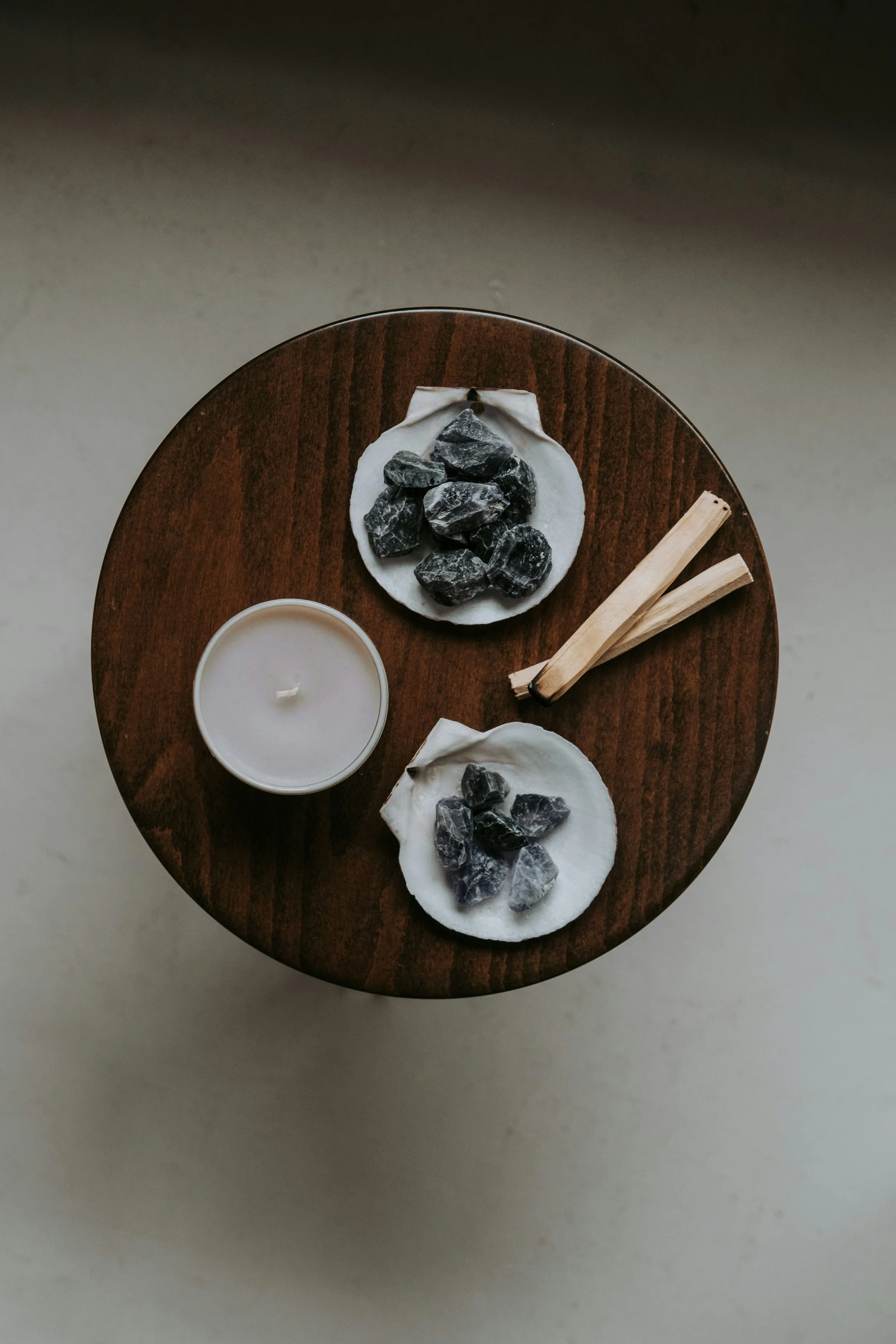 A round wooden table with two white coasters holding black stones, a small white candle, and two wooden sticks.