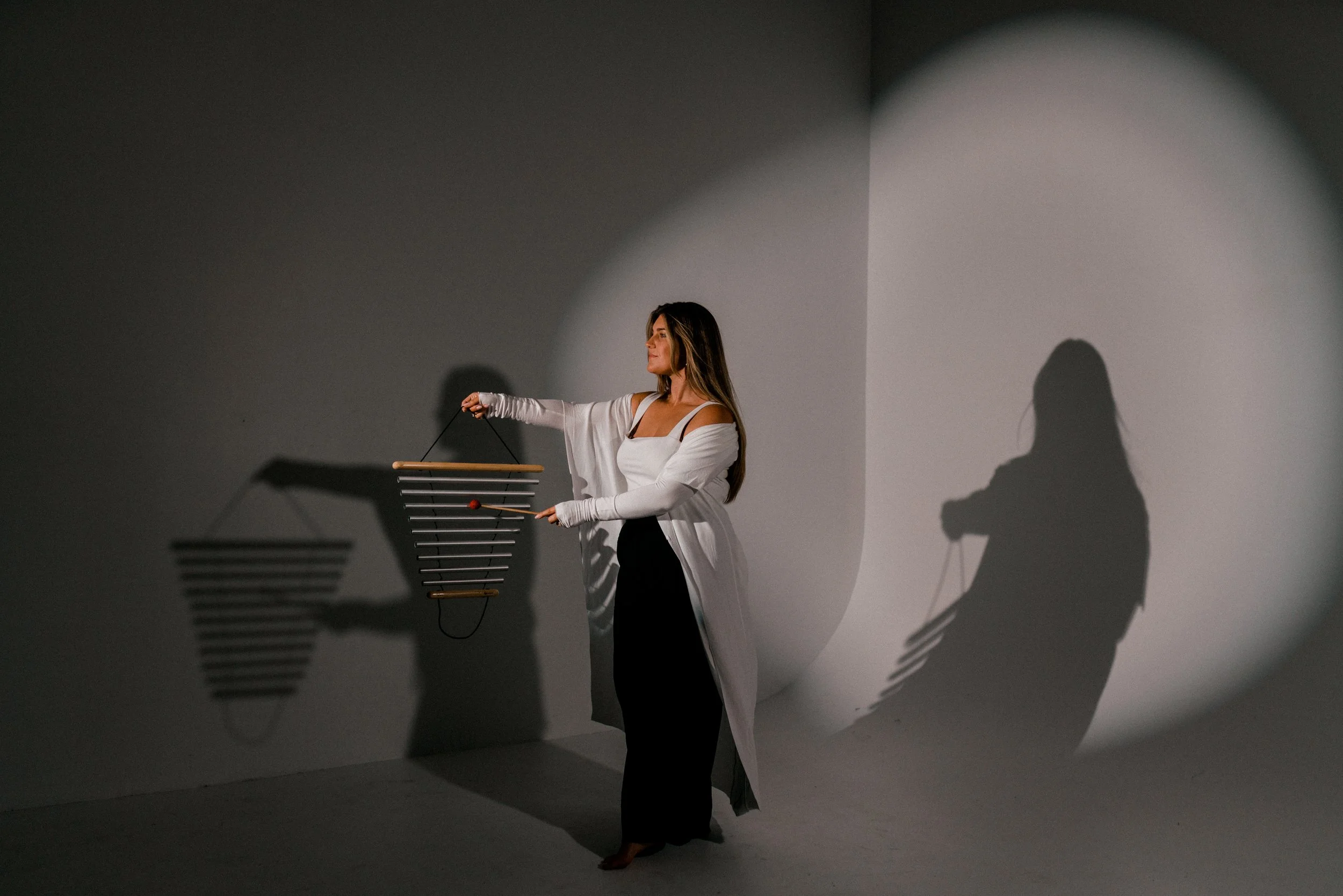 A woman in a white dress and long gloves holds a modern musical instrument made of horizontal black bars in front of a plain wall with spotlight shadows.