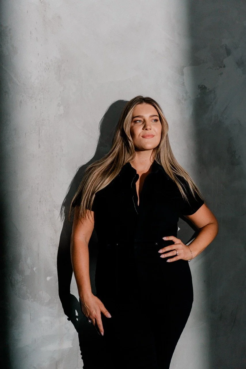 A woman with long blonde hair wearing a black outfit, standing with one hand on her hip, looking contented against a textured gray wall, with shadows cast on her face and body.