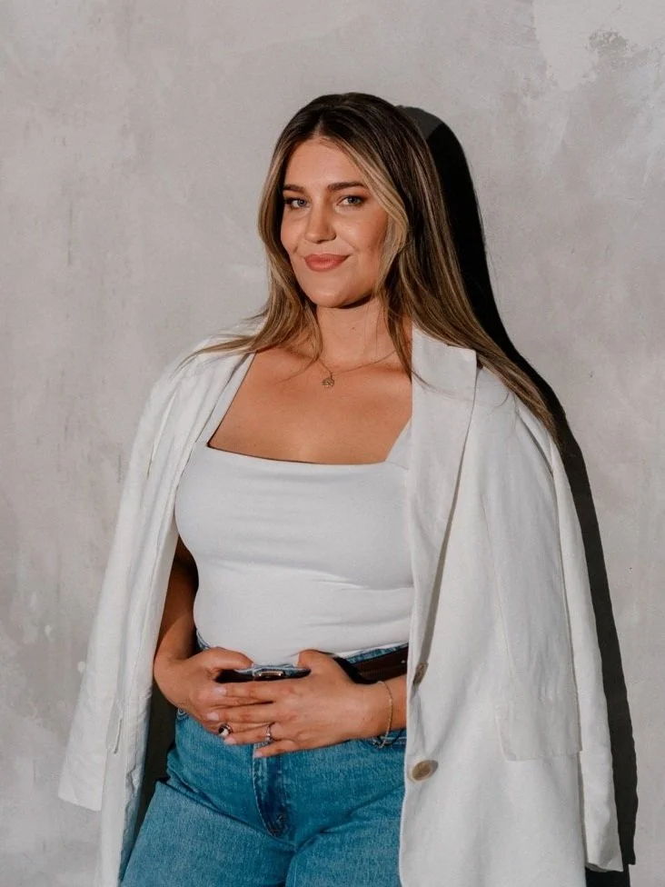 A young woman with long blond hair, wearing a white tank top, a white jacket draped over her shoulders, and blue jeans, standing against a plain gray wall, holding a smartphone, with a subtle smile.