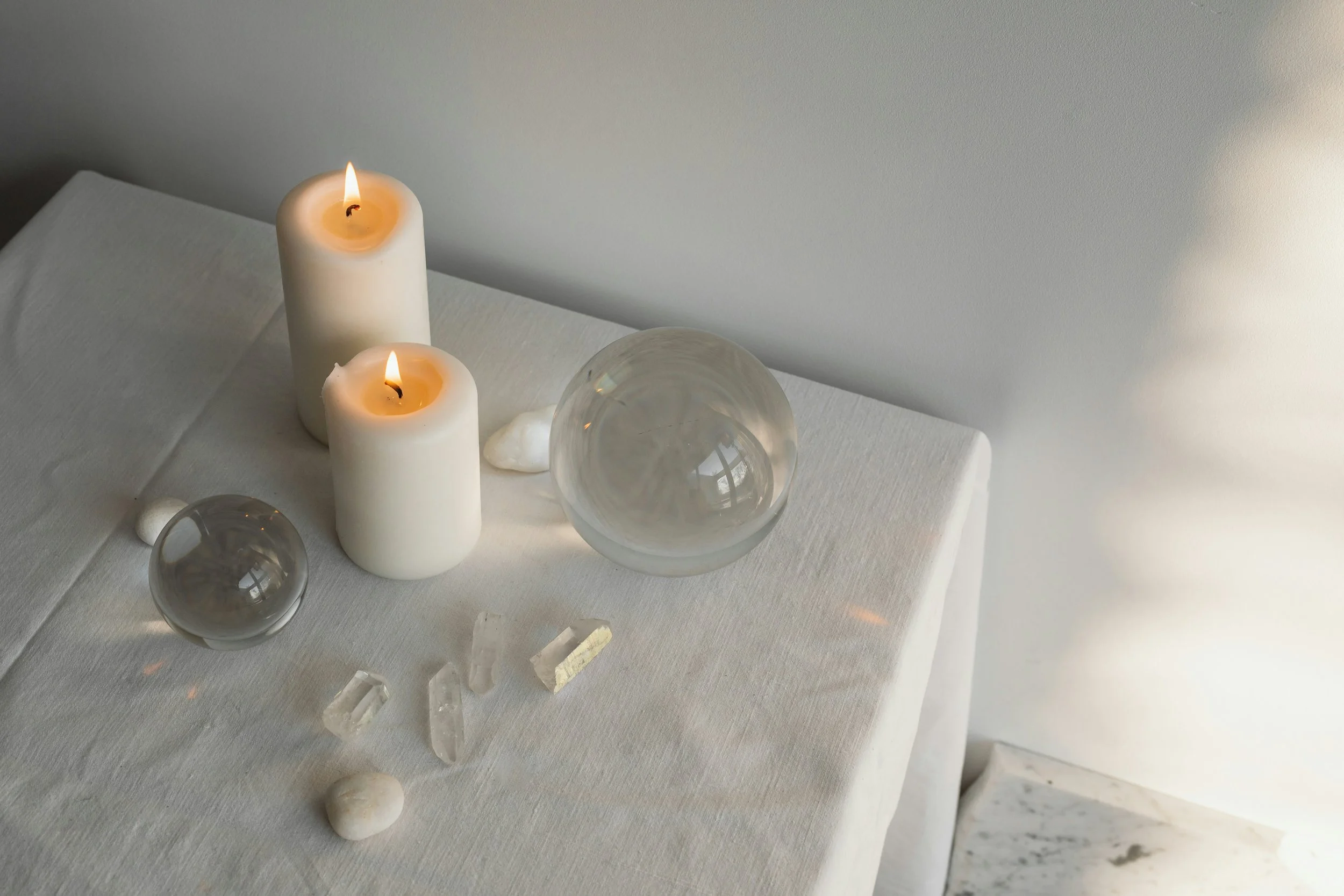 A white table with two lit white candles, two glass spheres, and various small rocks and crystals, set against a light gray wall background.