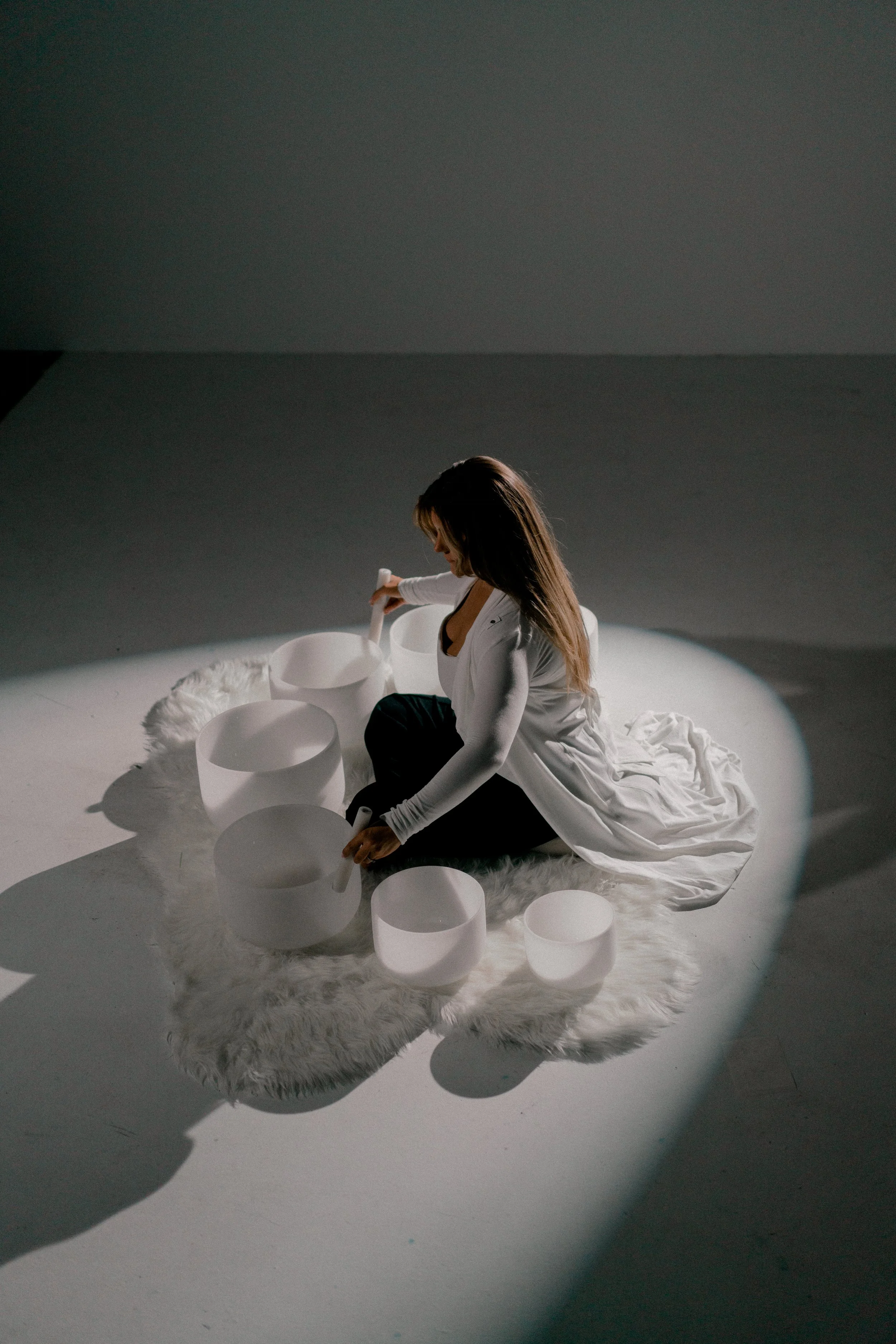A woman sitting on a fluffy white rug in a dark room, surrounded by large white singing bowls, illuminated by a spotlight.