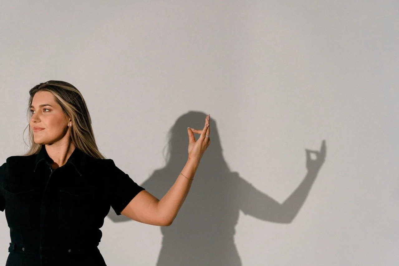 A woman in a black shirt making a gesture with her right hand, casting a shadow on a plain white wall that mimics her gesture.