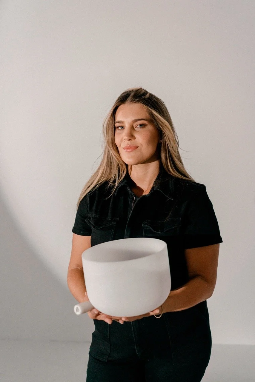 A young woman with long blonde hair and a confident smile, holding a white singing bowl with a mallet in front of a plain white wall.