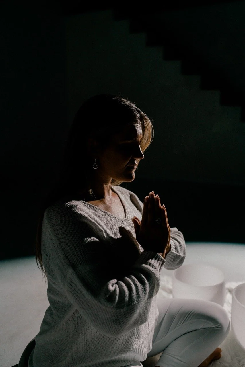 A woman is sitting cross-legged on the floor with her hands pressed together in a prayer position, possibly meditating, with dim lighting casting shadows on her face.