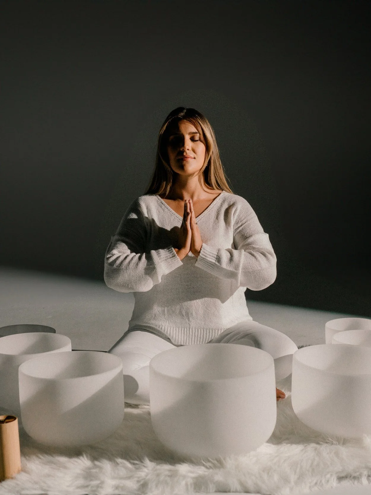 A woman sitting cross-legged on a fluffy white rug with her hands in prayer position, surrounded by multiple white singing bowls, on a neutral background.