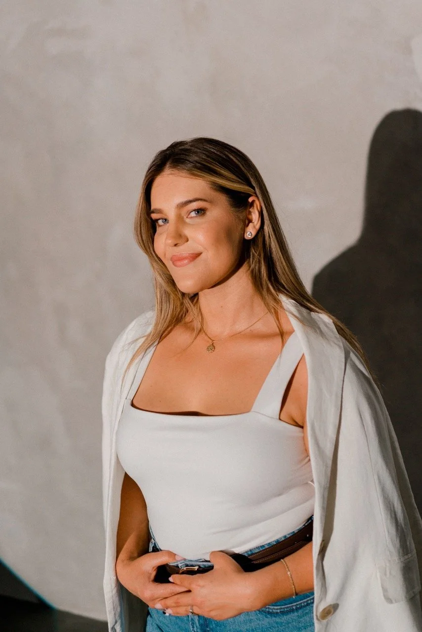 A woman with light brown hair smiling, wearing a white tank top, a white jacket draped over her shoulders, blue jeans, and jewelry, standing against a plain background.