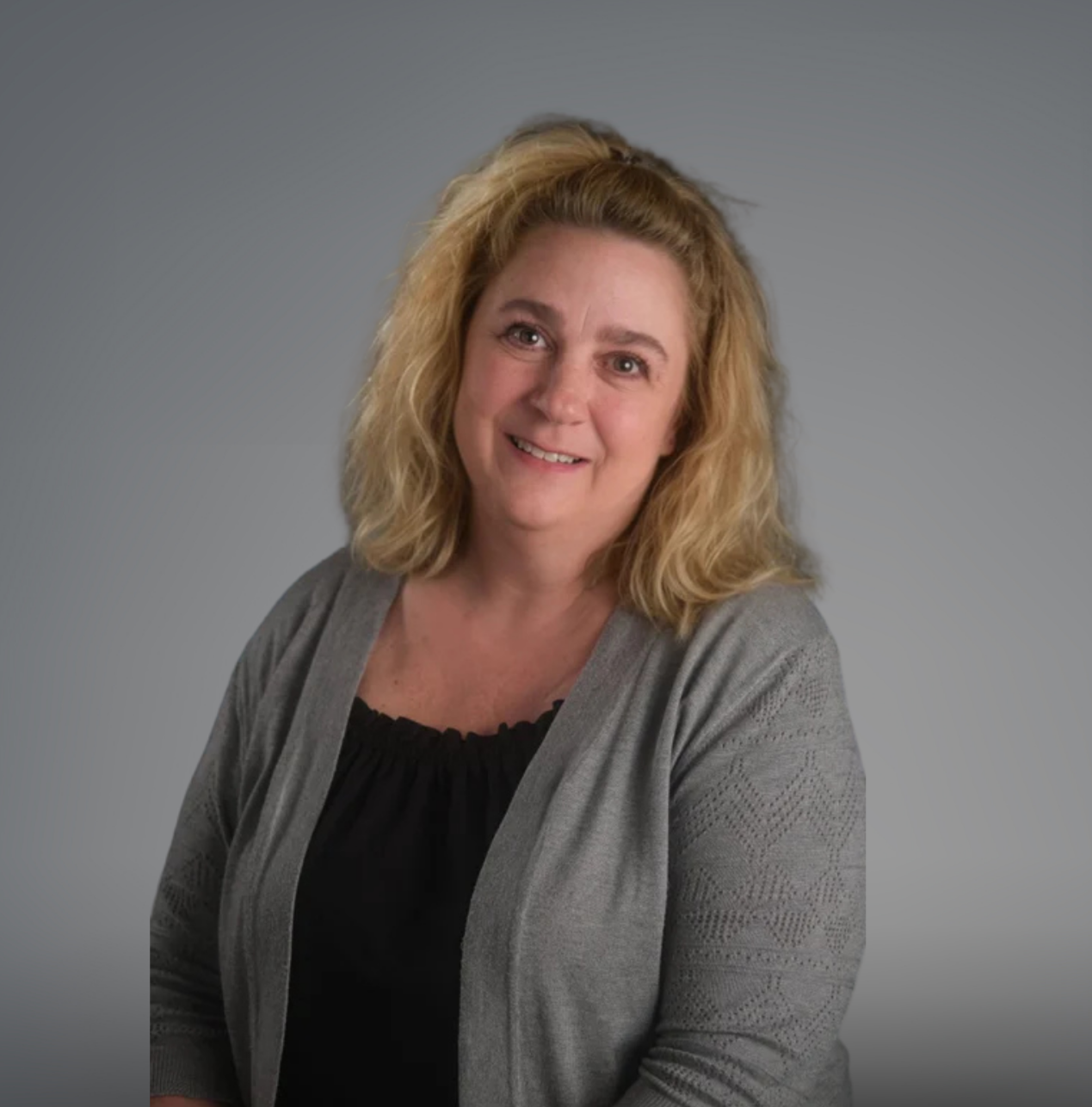 A woman with blonde, shoulder-length, curly hair smiling at the camera against a dark, textured background. She is wearing a black top with a ruffled neckline and a light gray cardigan with a subtle geometric pattern.