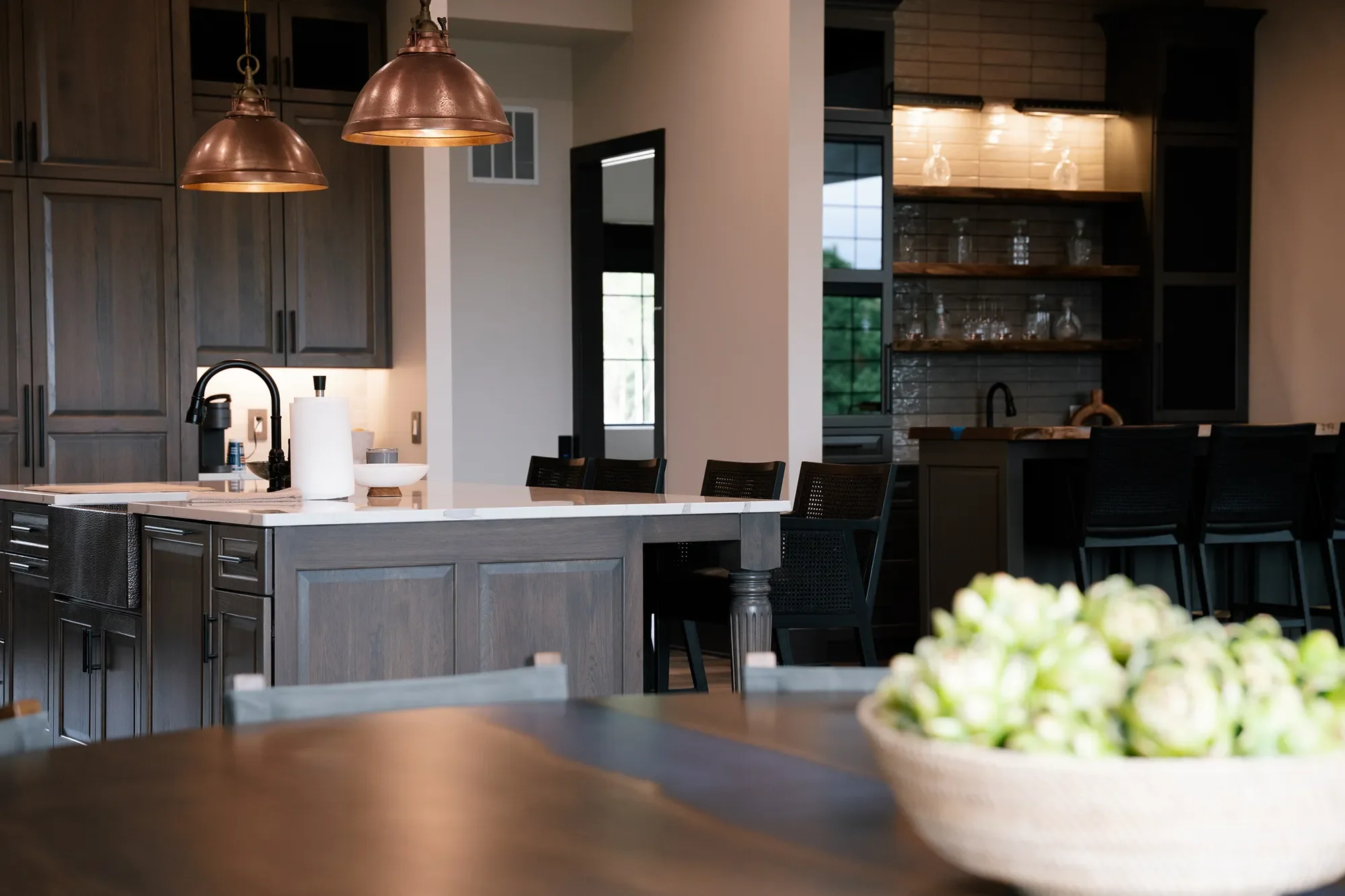 Modern kitchen with dark wood cabinets, an island with a white marble countertop, two copper pendant lights, black chairs, and open shelving with glassware.