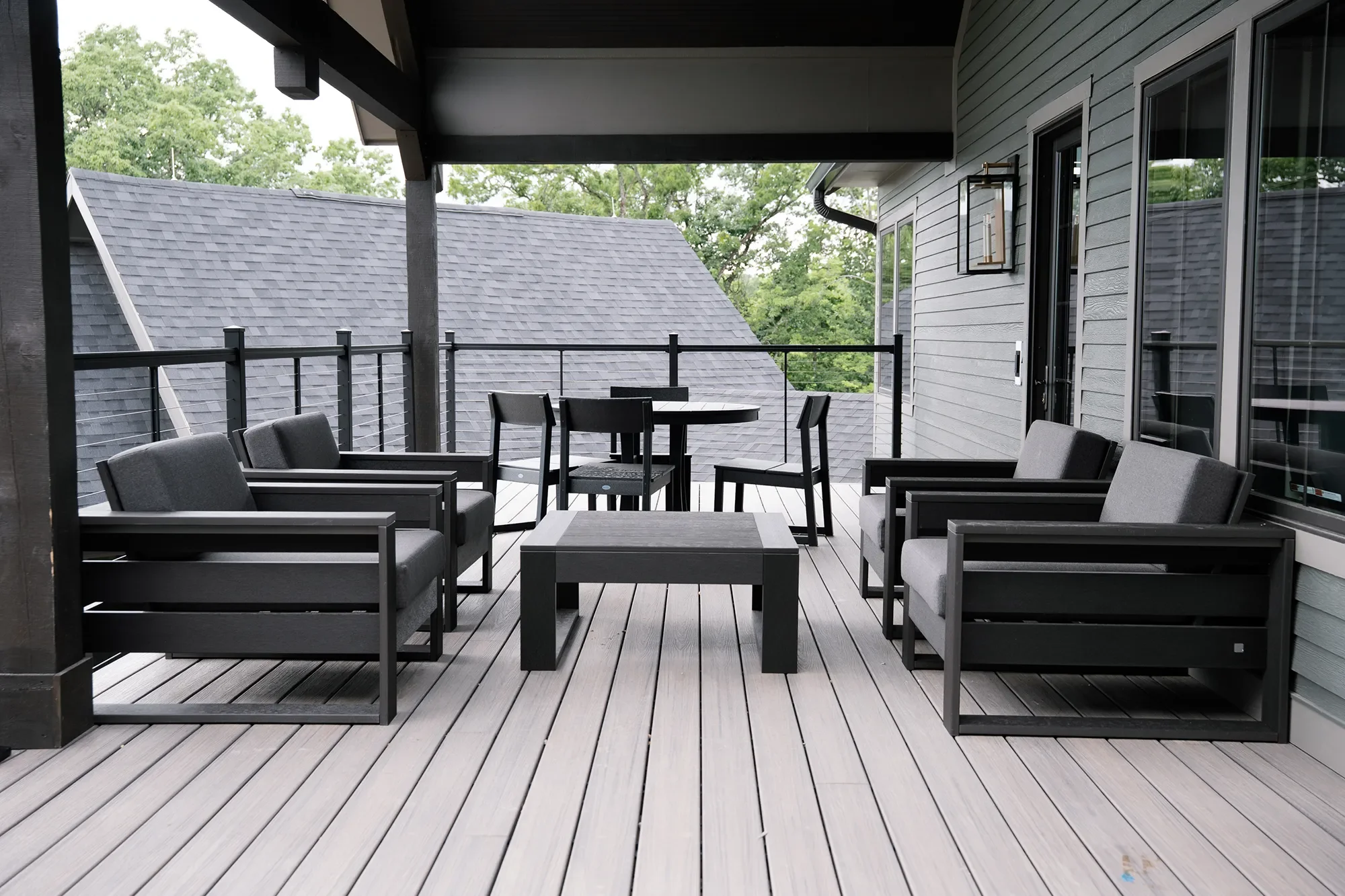 View of a modern balcony with outdoor seating including gray armchairs, a small black table, and a round dining table with black chairs. The balcony has gray wood decking, black railing, and is attached to a gray house with horizontal siding. Tree br