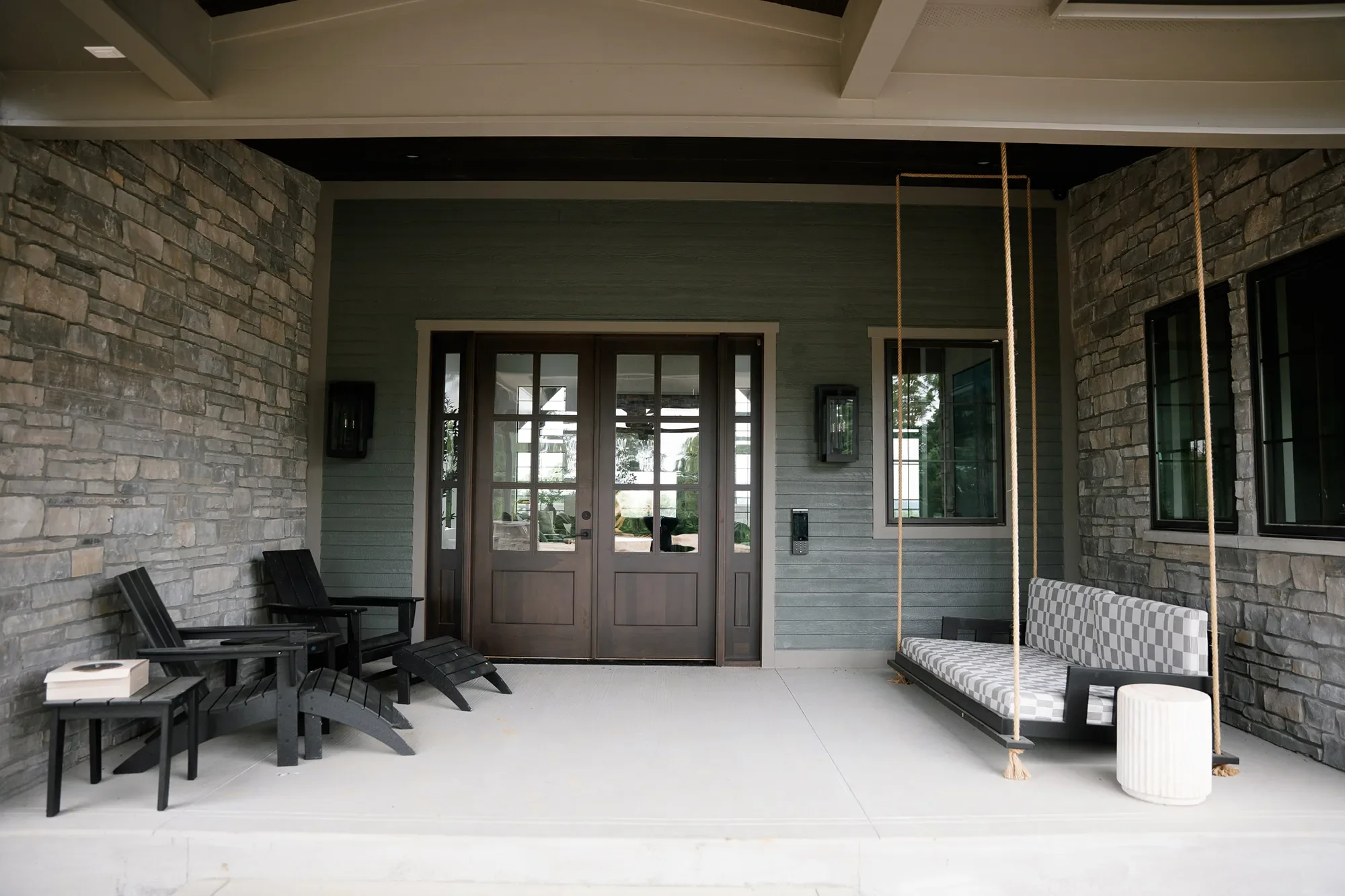 Covered porch with black chairs, a bench with a checkered cushion, a swing with a rope, and stone and siding walls with windows and door.