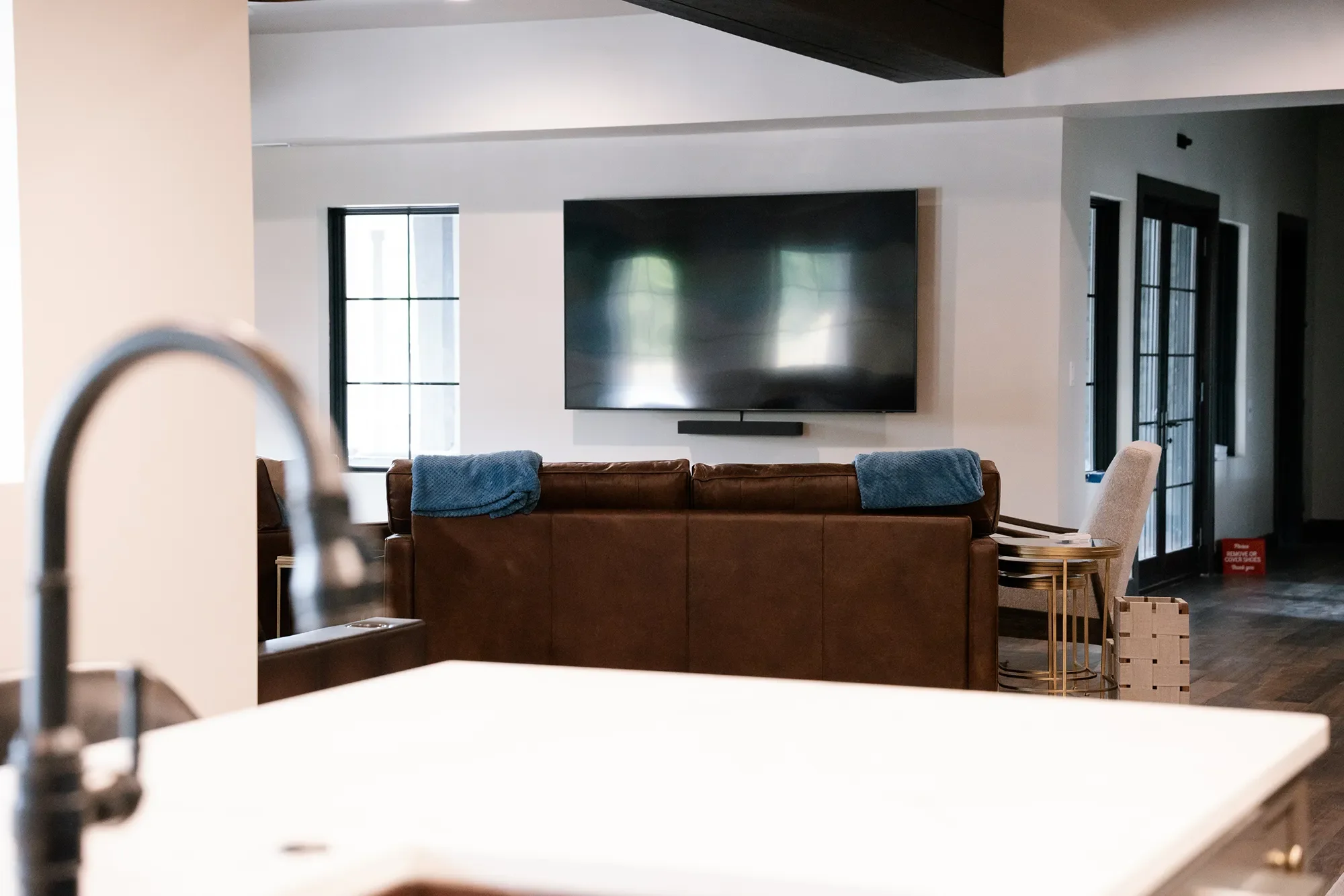 Living room with a large flat-screen TV mounted on the wall, a brown sofa with blue towels laid on it, and windows with black frames. Part of a dining chair and a small side table are visible.
