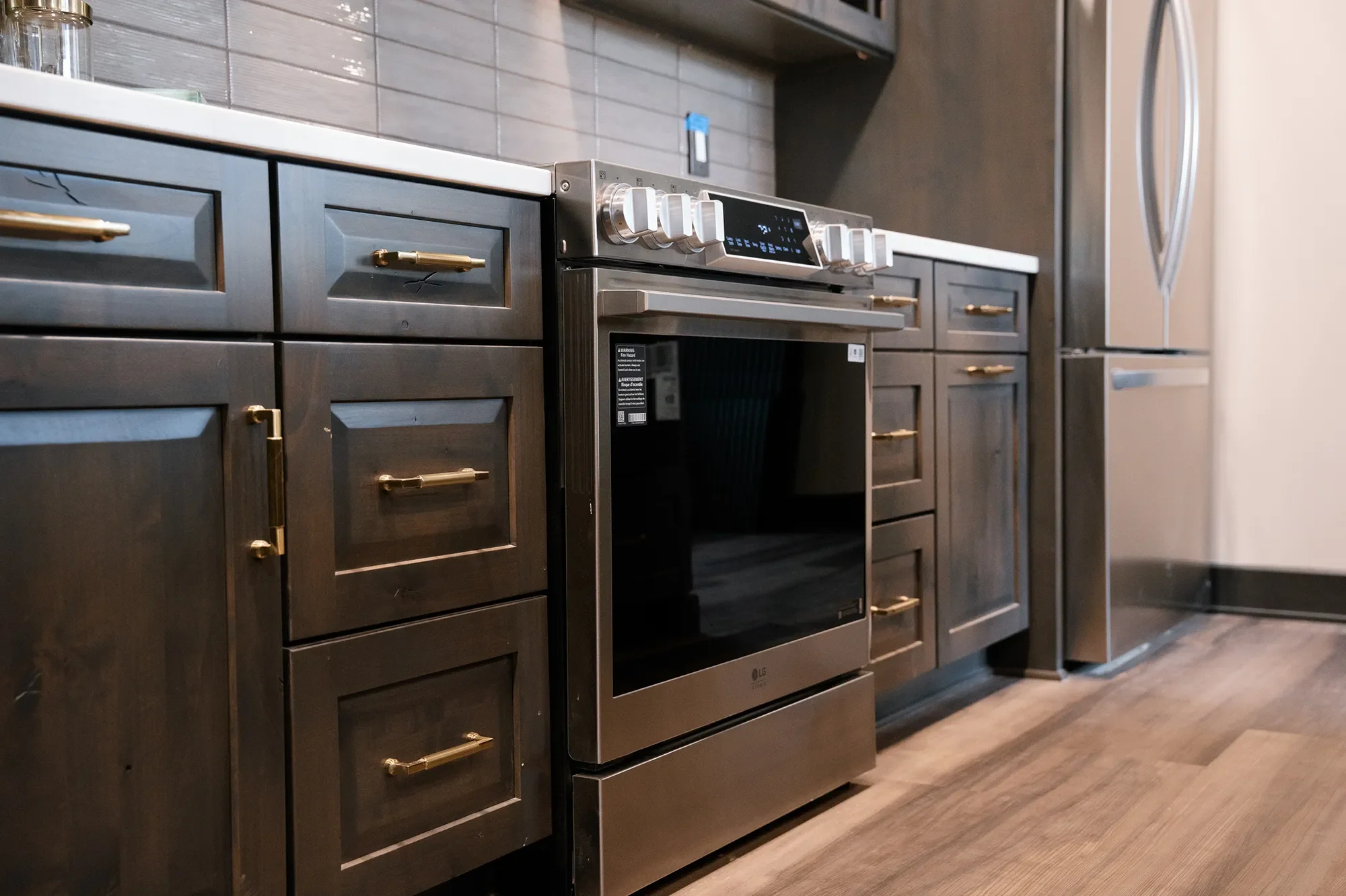 A modern kitchen with dark cabinetry, gold handles, a stainless steel oven, and a gray tiled backsplash.