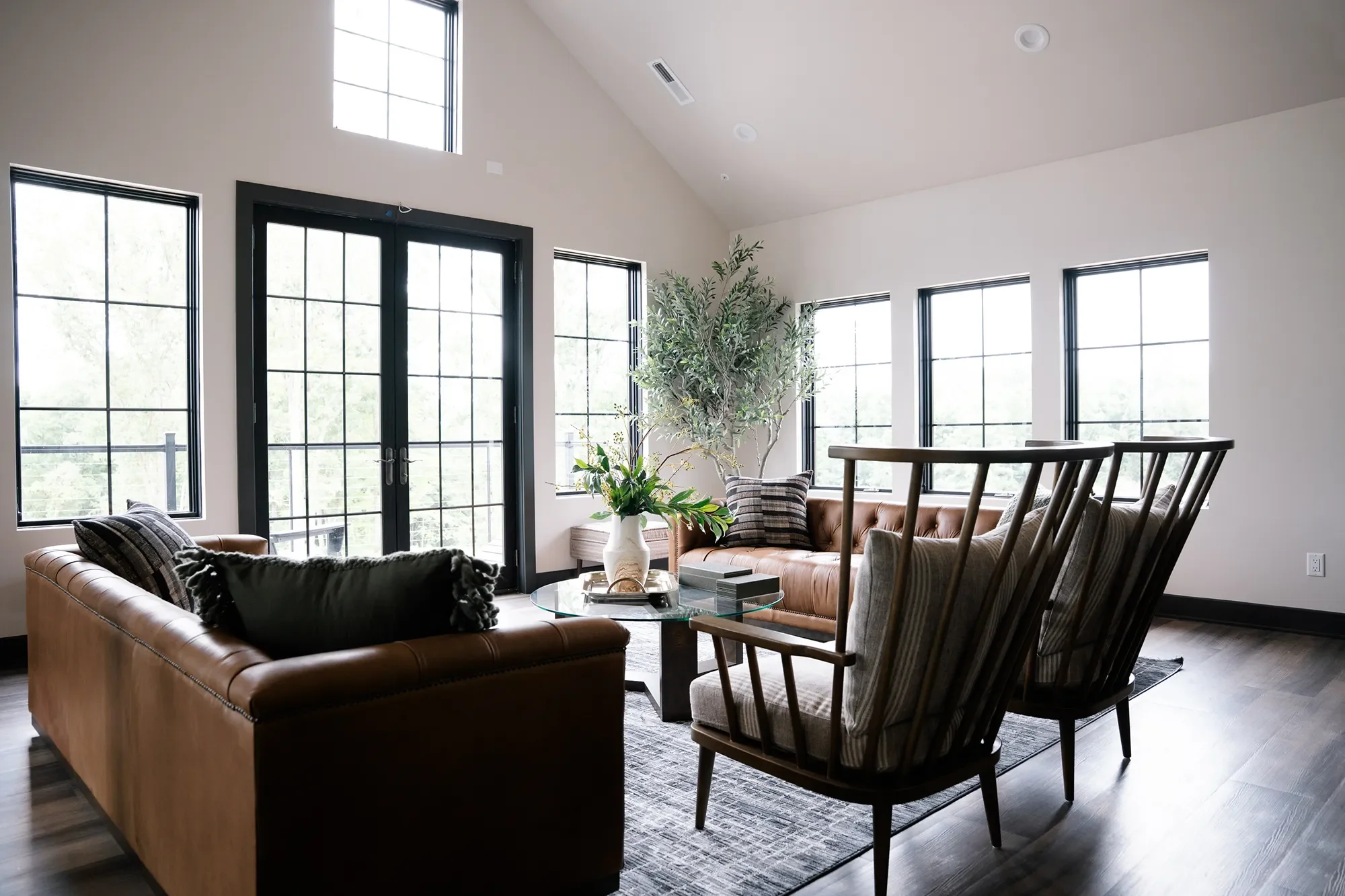 Bright living room with large windows and glass door, beige leather sofa, wooden chairs, a glass coffee table with books and a plant, and a tall potted plant in the corner.