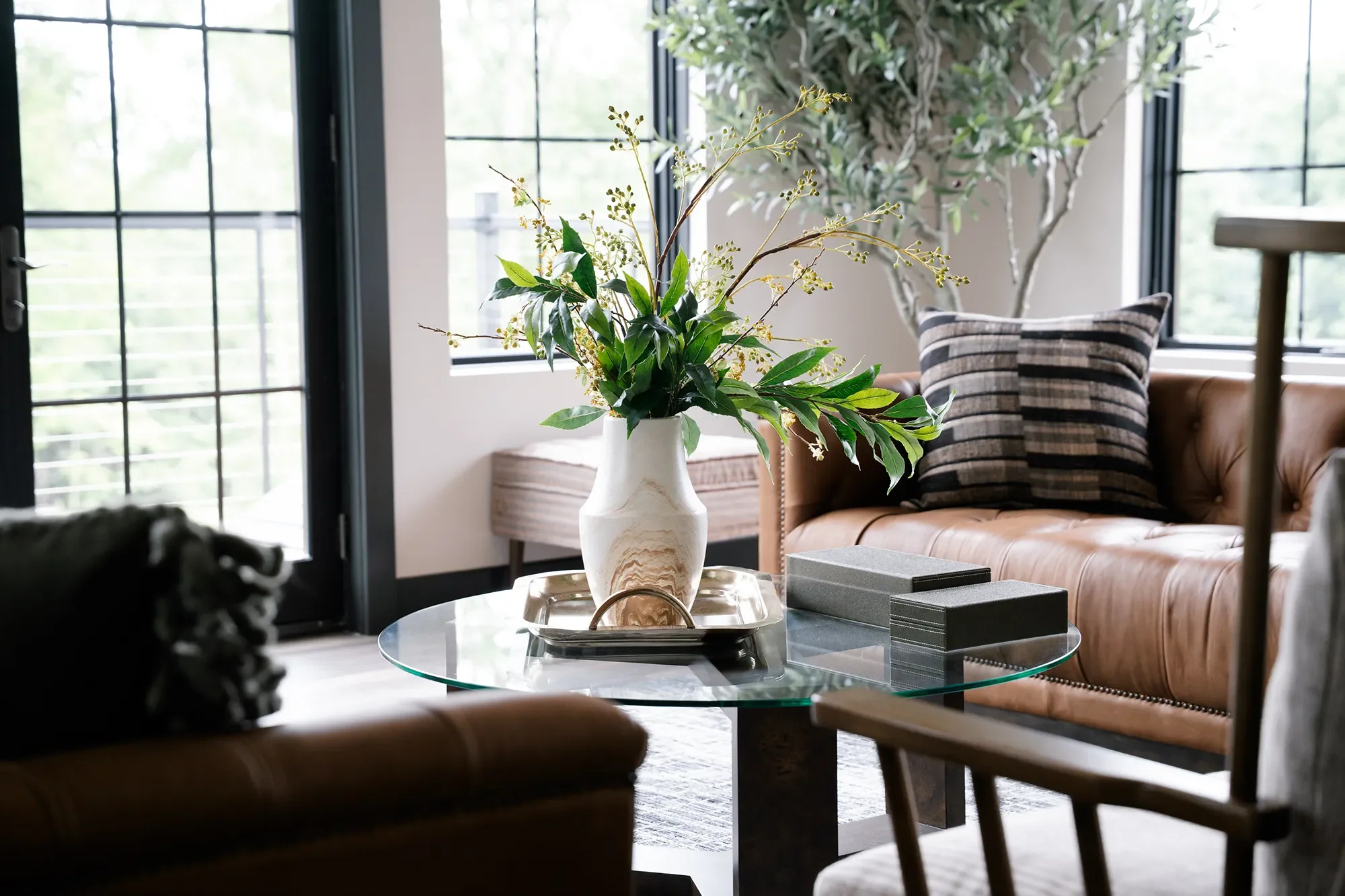 Living room with large windows, beige leather sofa with striped cushion, glass coffee table with a vase of green leafy plant, books, and decorative tray, and a potted tree in the background.