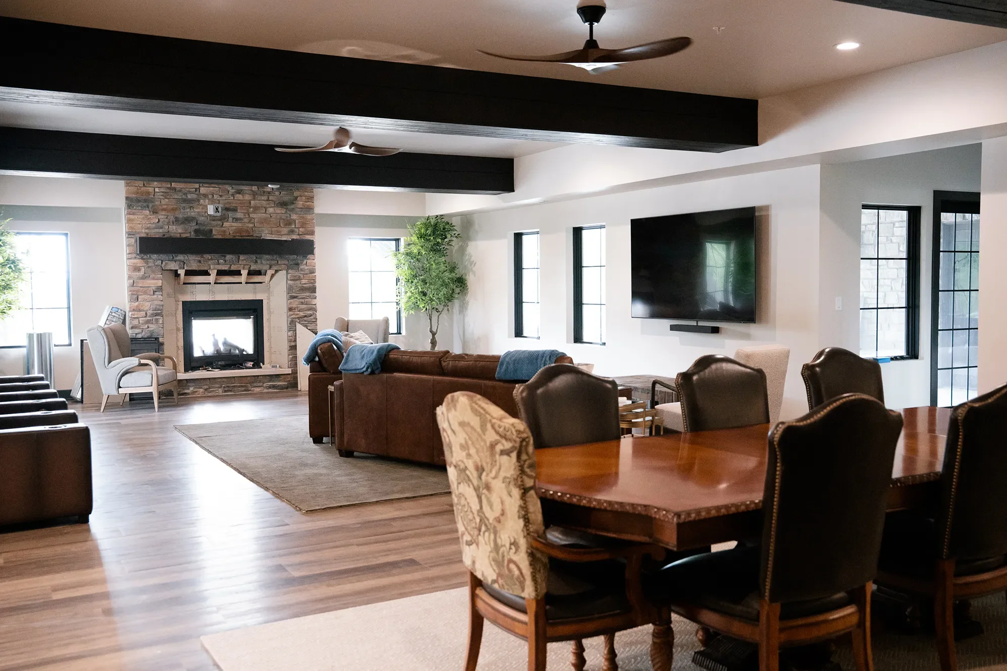 Living room with a stone fireplace, a large flat-screen TV, multiple windows, ceiling fans, and various chairs and couches.