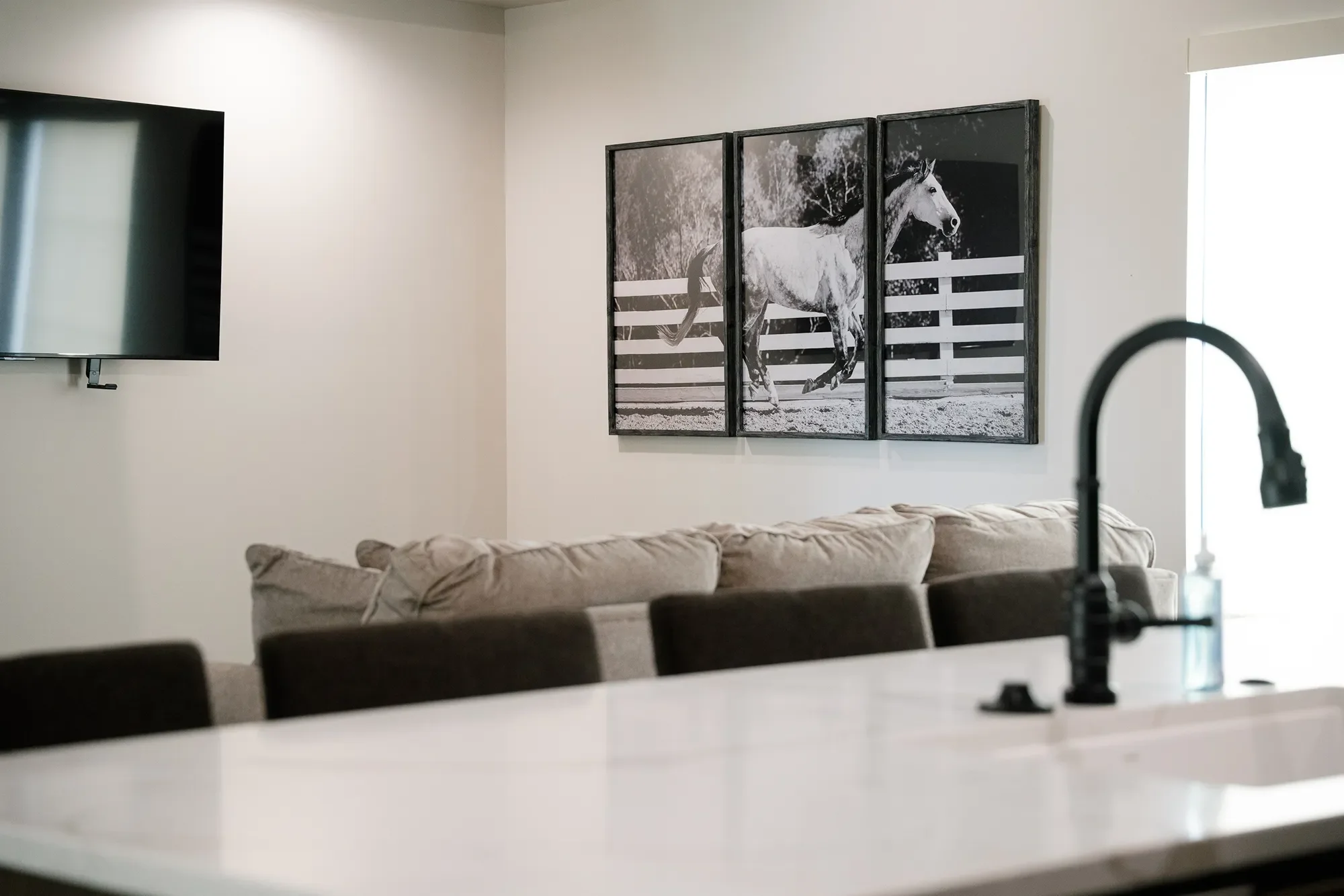 Living room with a triptych black and white painting of a running horse on the wall, beige couch below it, dark chairs around a white table, flat-screen TV mounted on the wall, and a kitchen faucet visible in the foreground.