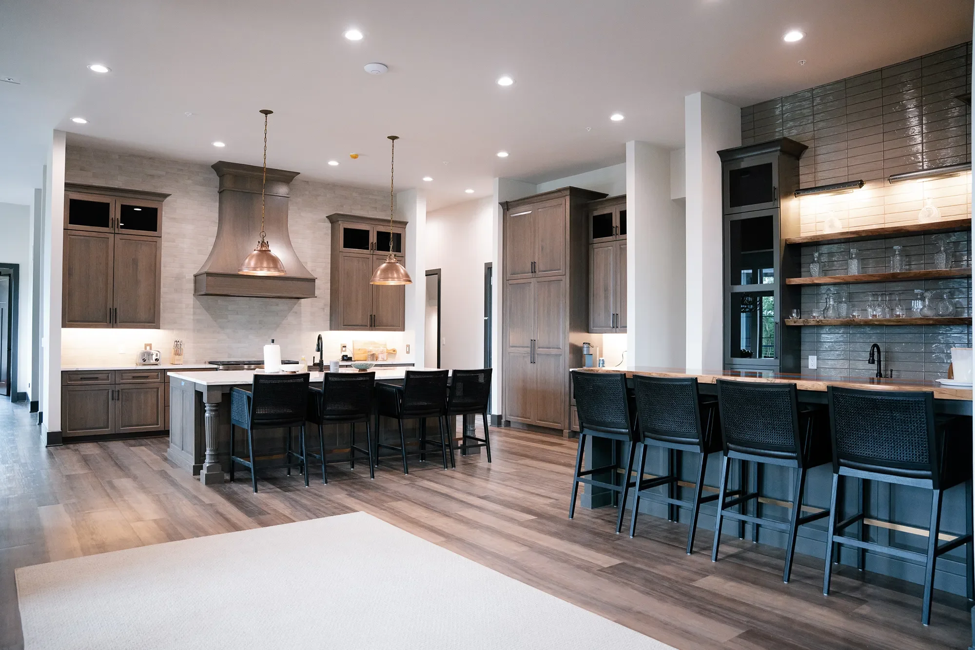 Modern kitchen with wooden cabinets, a large kitchen island with black chairs, pendant lights, and open shelving on a gray brick wall.