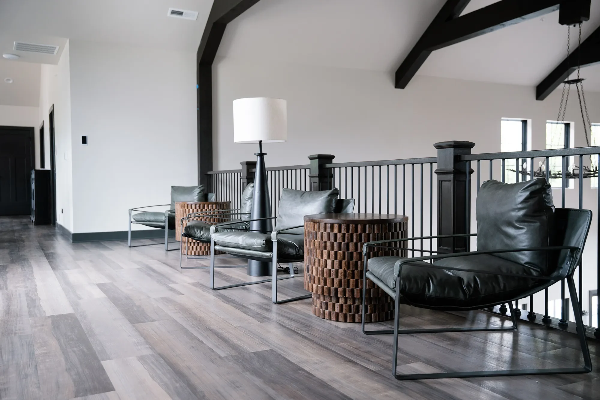 Modern living room with black leather chairs, wooden side tables, a tall black floor lamp, and a black metal railing. The room features a vaulted ceiling with dark exposed beams, small rectangular windows, and wood flooring.
