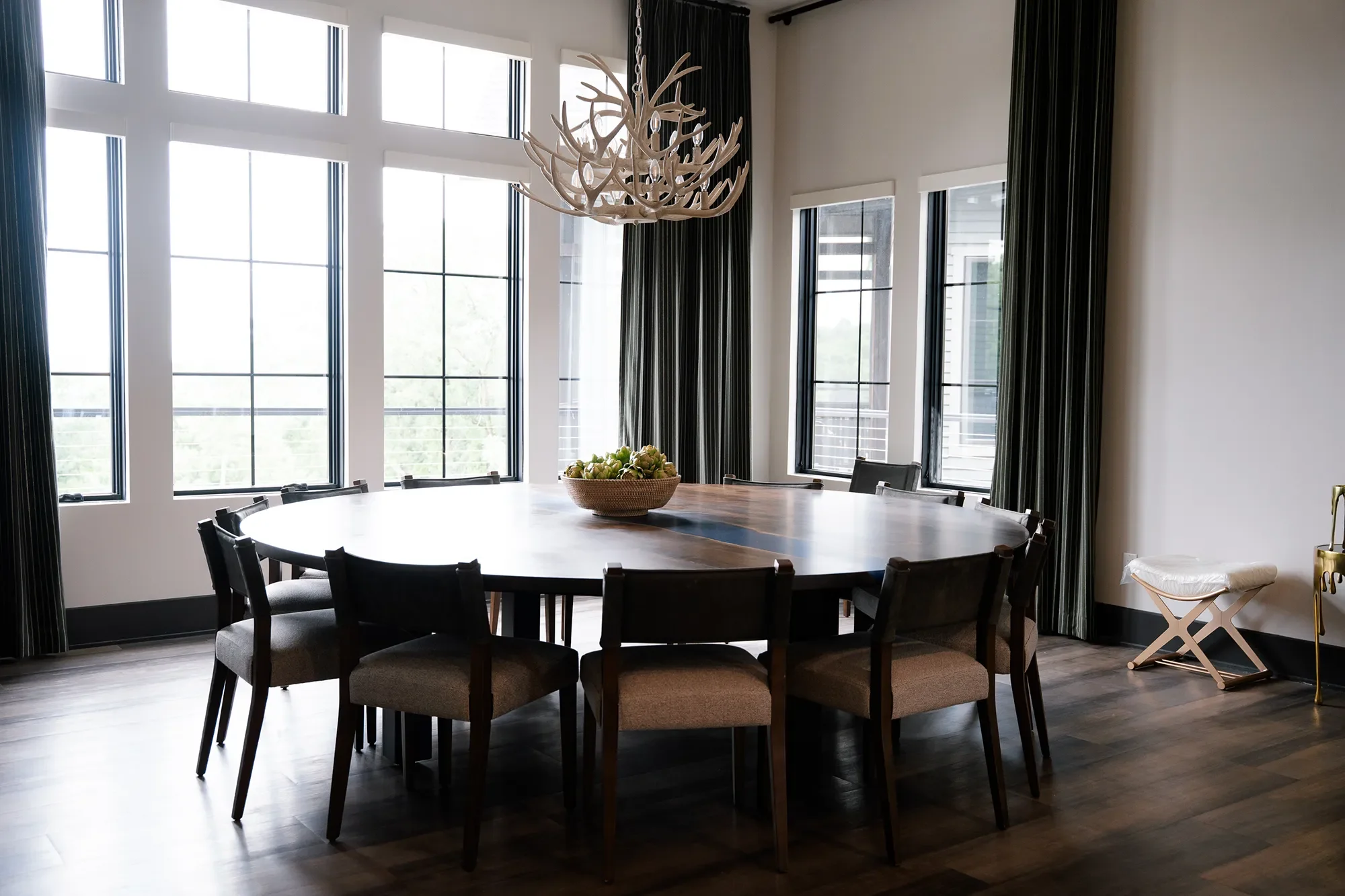 A modern dining room with a large wooden table surrounded by eight dark upholstered chairs, a decorative bowl of green apples in the center, large windows with black frames letting in natural light, black curtains, a hanging antler chandelier, and a 