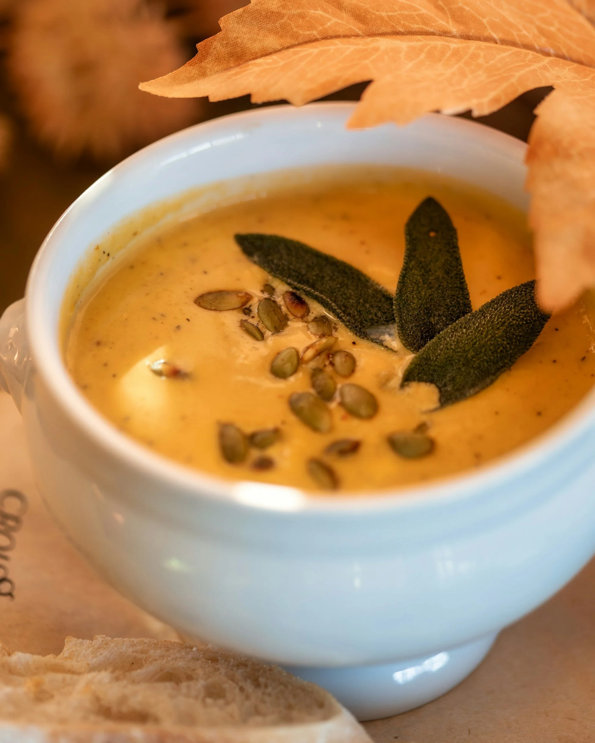 A white cup filled with pumpkin soup garnished with sage leaves and pumpkin seeds, with a piece of bread nearby and autumn leaves in the background.