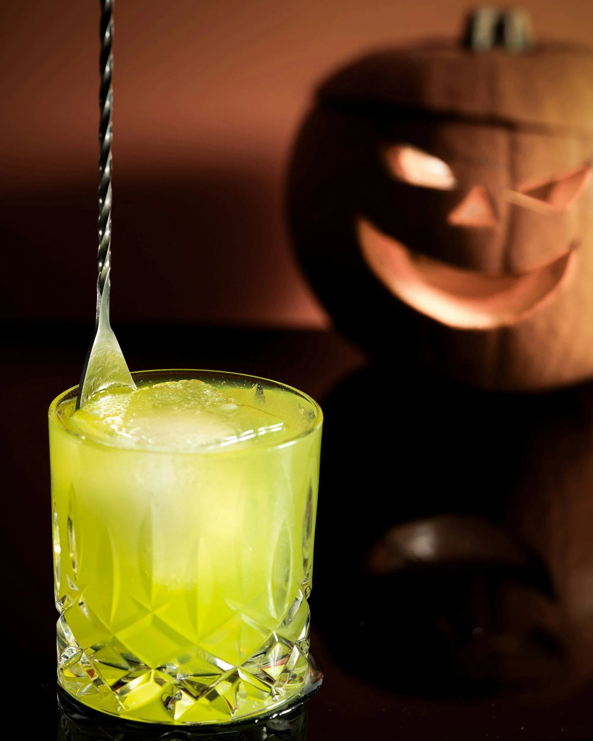 A green cocktail in a cut-glass tumbler with a long twisted metal spoon, set on a reflective black surface, with a carved wooden pumpkin featuring a smiling face in the background.