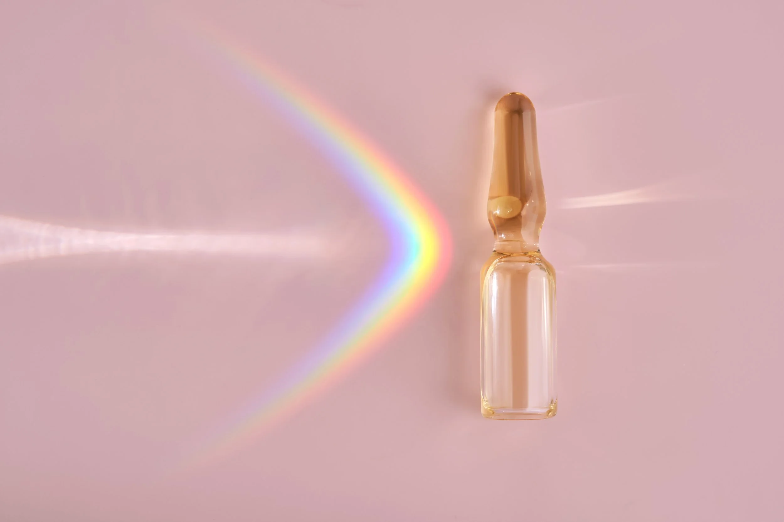 A glass ampoule with a clear liquid inside on a light pink background, with a rainbow reflection to the left.