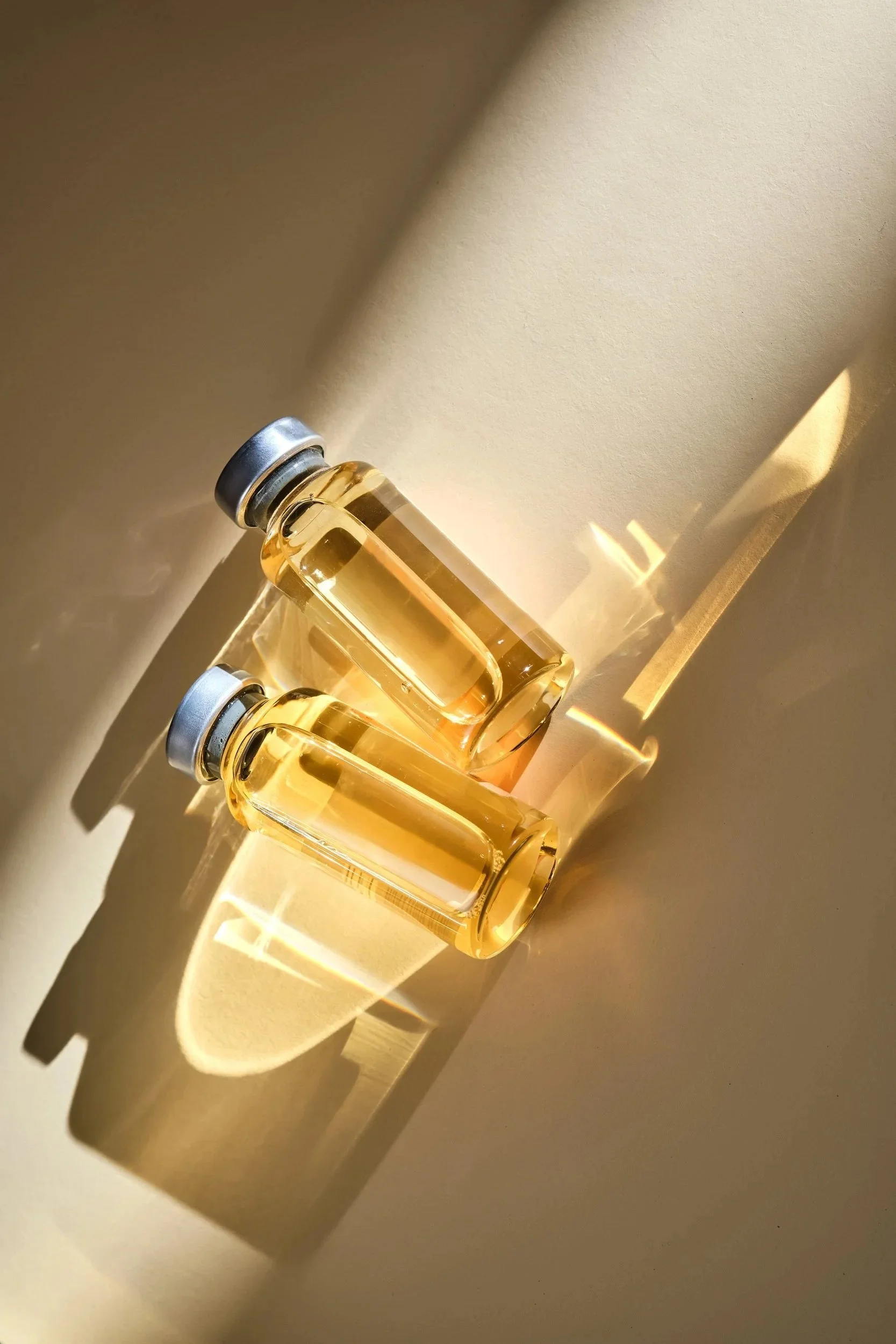 Two small glass bottles filled with golden liquid, placed on a beige surface with sunlight casting shadows and reflections.