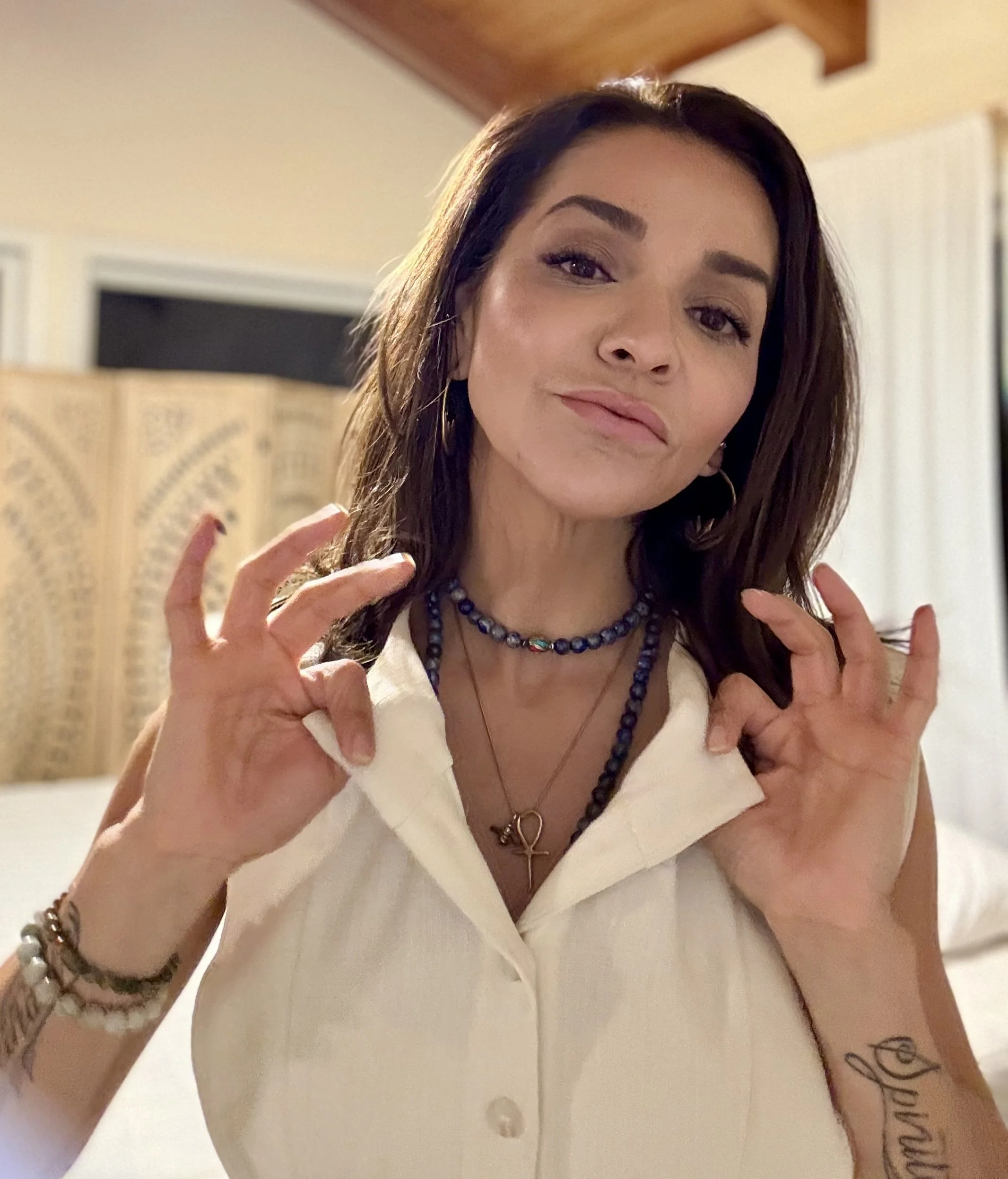 A woman with dark brown hair, wearing a white sleeveless shirt, multiple necklaces including an ankh pendant, and bracelets, making a peace sign with both hands and slightly smiling.