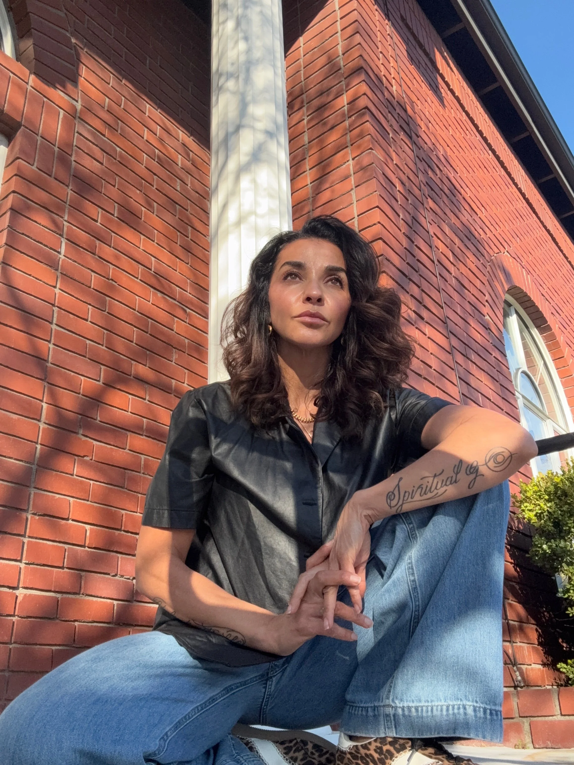 A woman with dark curly hair, wearing a black leather shirt and blue jeans, is sitting on the ground outside a brick building with arched windows. The sunlight casts shadows on the building.